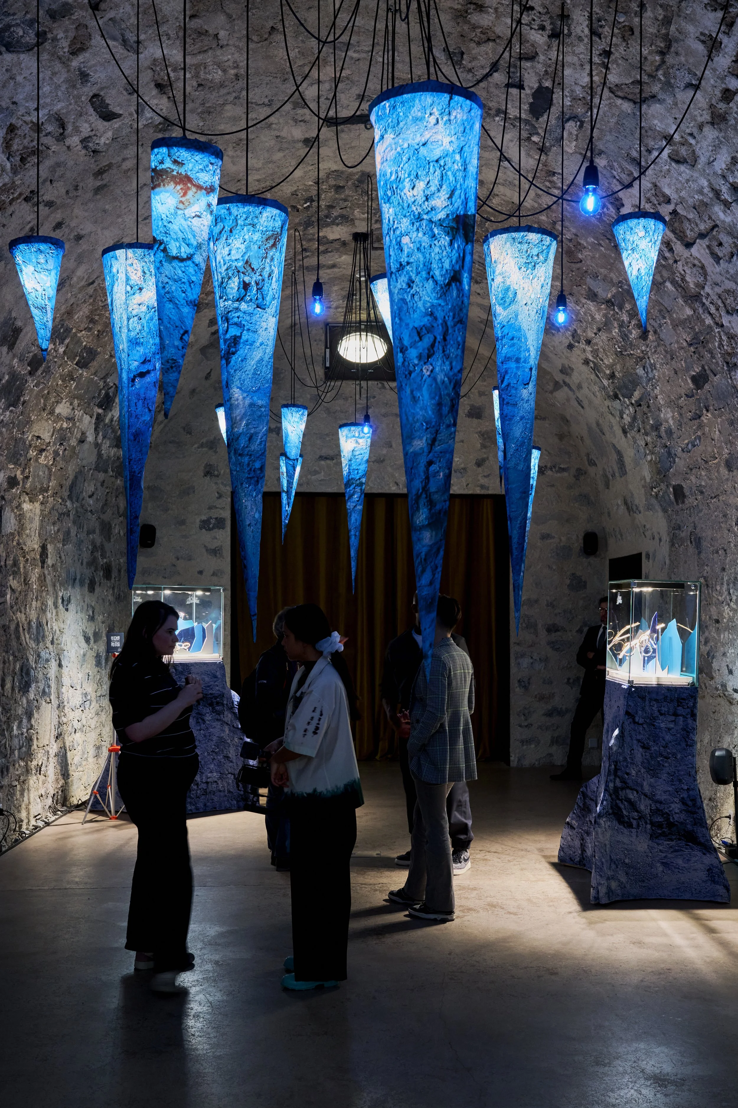 Décor de lampes suspendues en forme de cône, éclairage bleu, et des personnes discutant dans une galerie ou un musée avec mur en pierre.