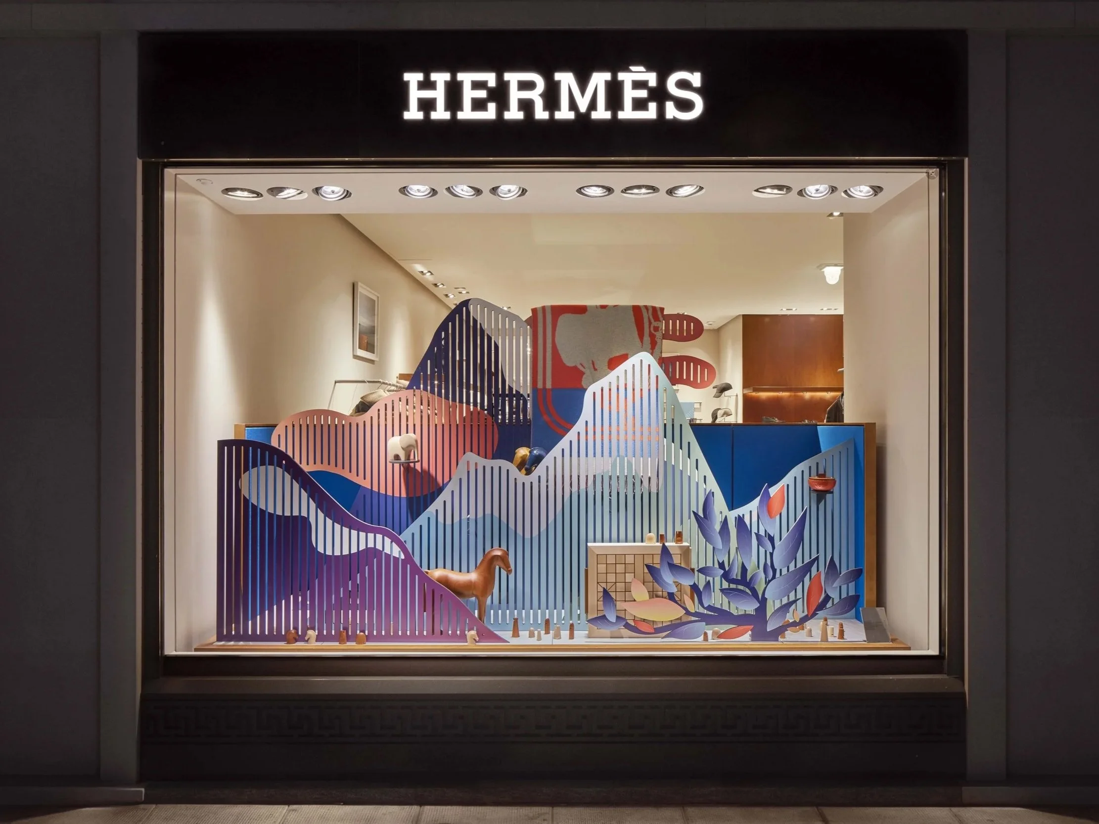 Vitrine d'une boutique Hermès avec scénographie colorée inspirée des paysage de montagnes suisses, mettant en lumières des objets Hermès : un plaid, un jeu d'échec, des sculptures en bois et des accessoires de décoration pour la maison.