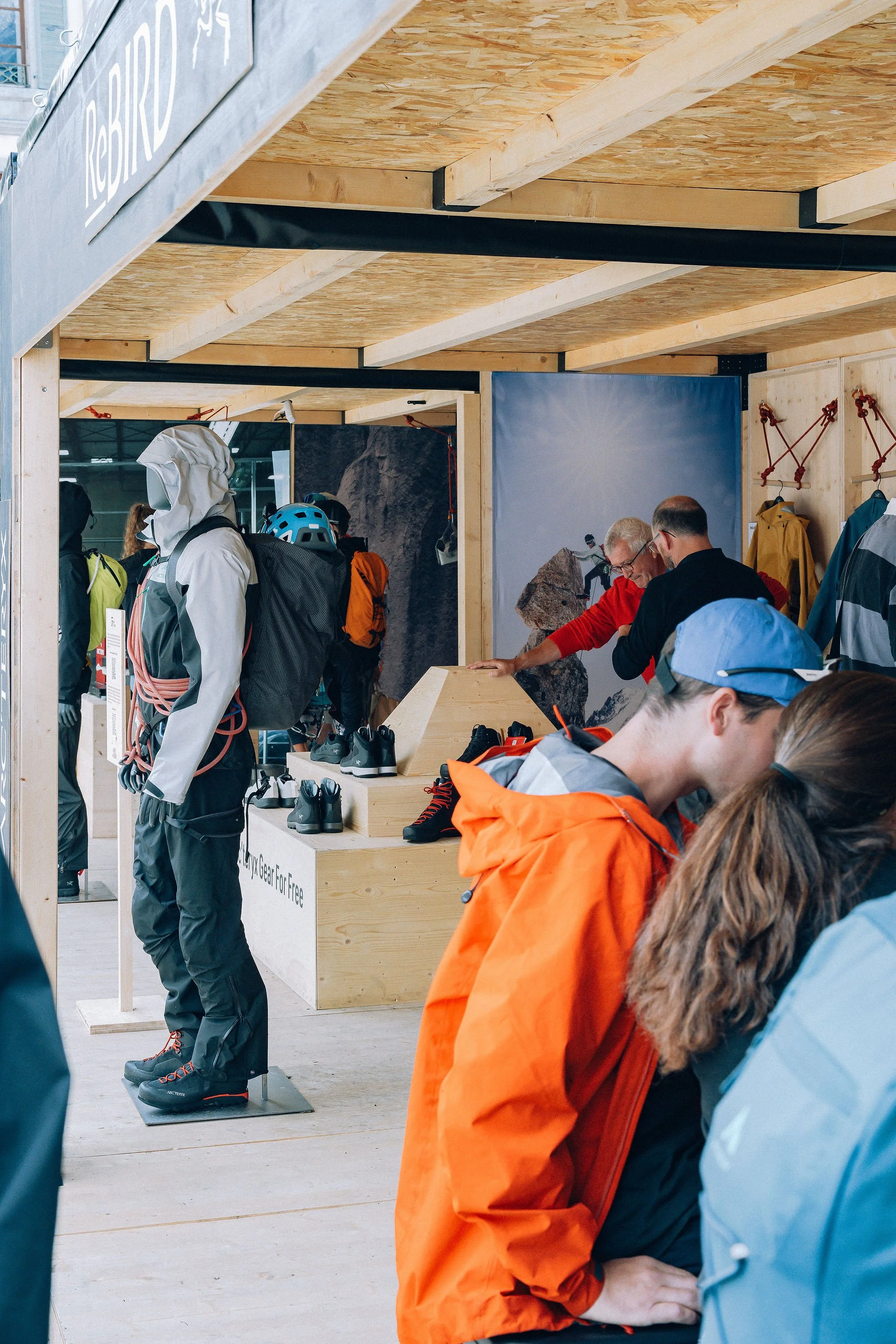 Groupe de personnes dans une exposition de matériel de montagne. Un homme en veste orange essaie des chaussures d'escalade, autres visitent et discutent dans cet espace.
