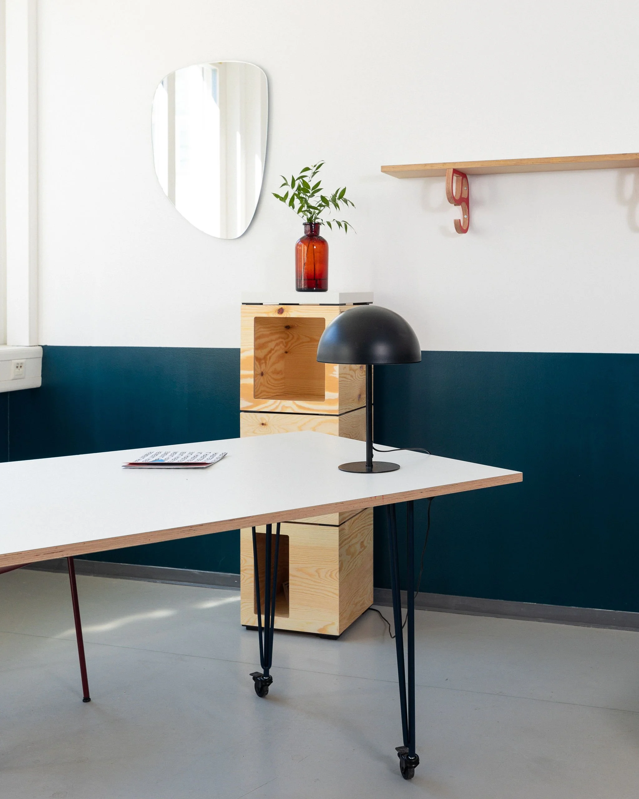 Une pièce moderne avec une table blanche, un lampadaire noir, une étagère en bois, une boite en bois, un miroir oval et un vase en verre avec des branches vertes.