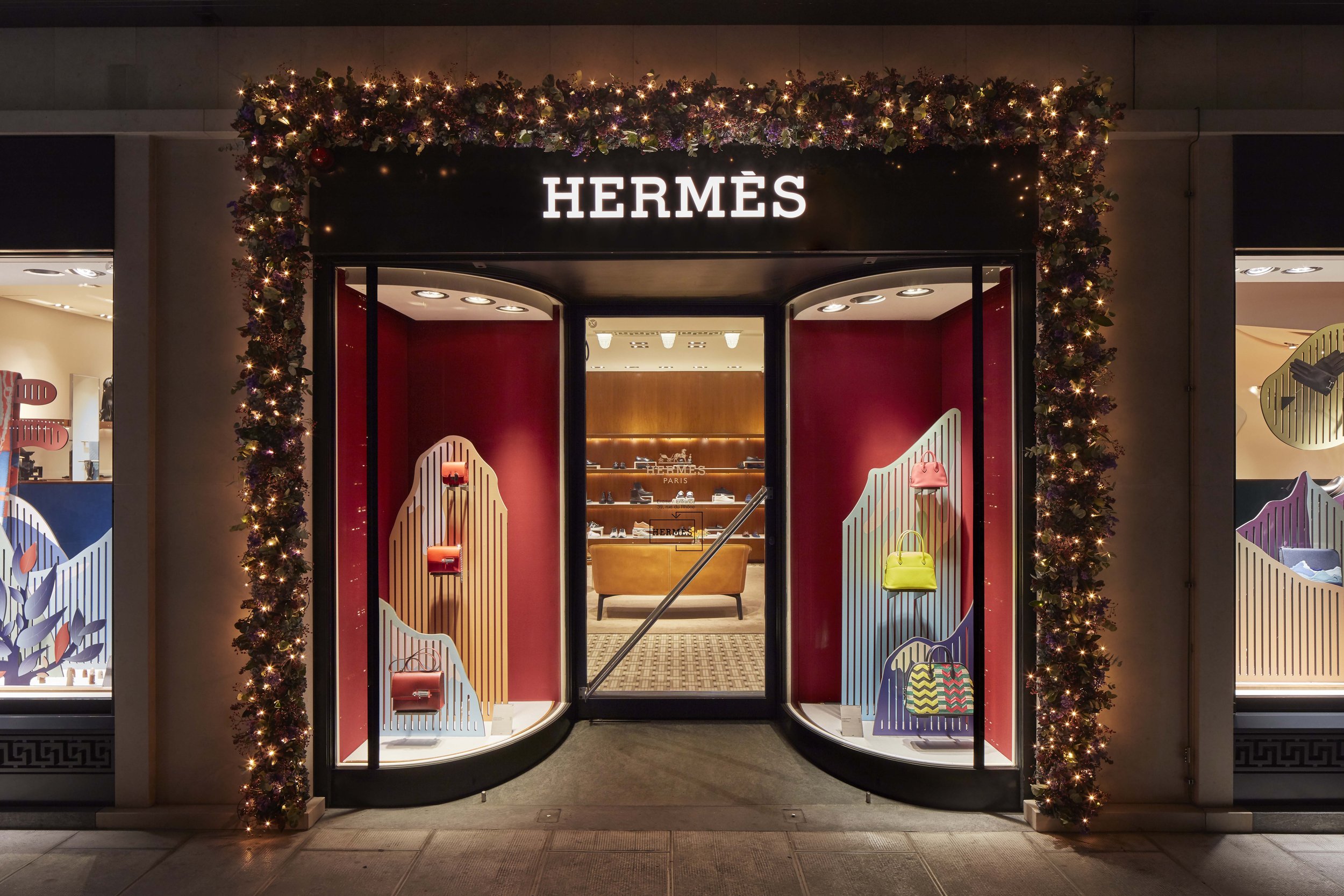 Vitrine dans l'entrée du magasin Hermès à Genève, décorée de guirlandes de Noël, avec des sacs à main colorés à l'intérieur mis en valeur par une scénographie contemporaine et colorée.