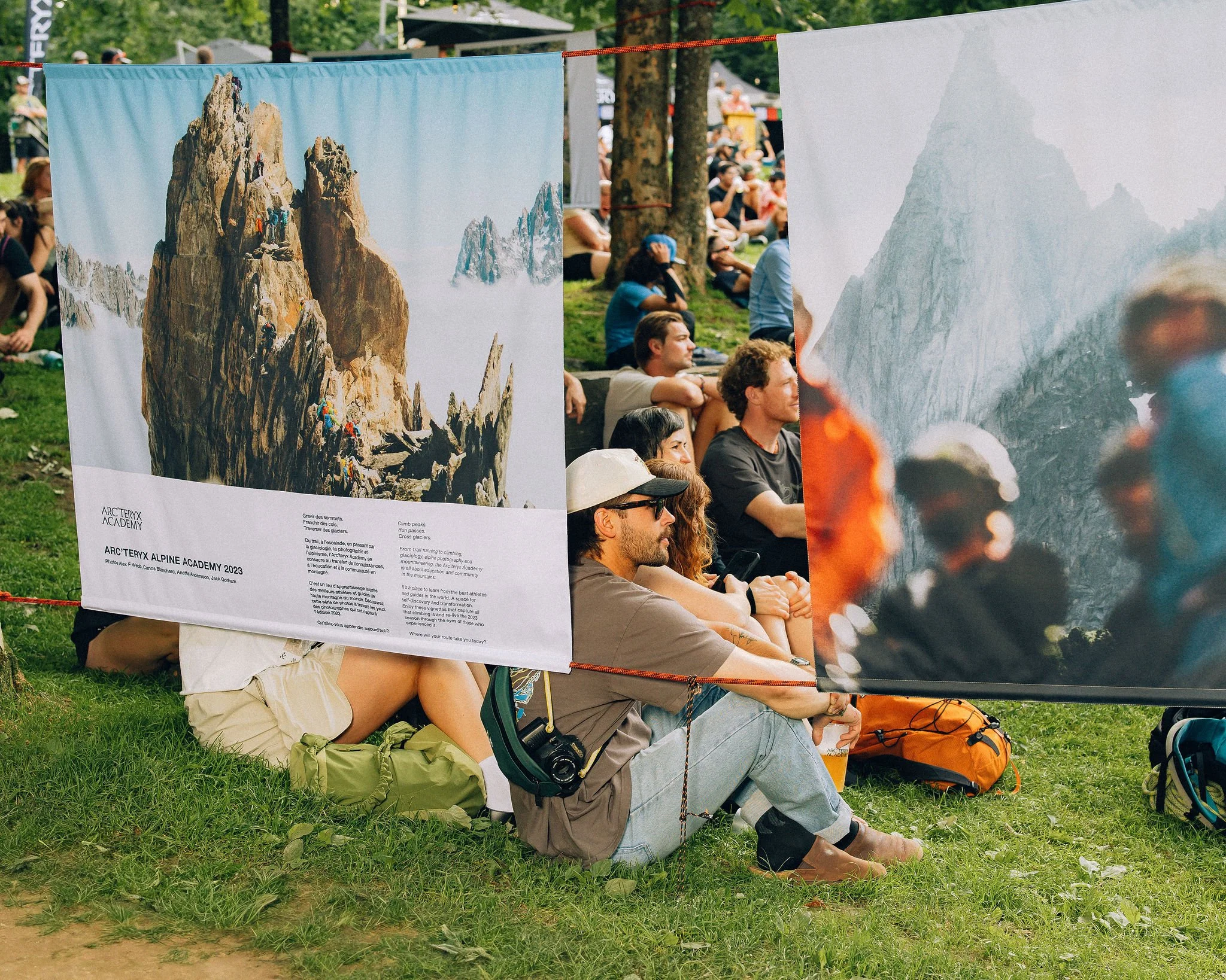 Affiches promotionnelles dans un parc avec des personnes assises sur l'herbe, regardant les affiches. L'une montre une image d'une montagne avec des grimpeurs. L'autre est floue, mais semble aussi représenter une montagne. Le groupe est détendu dans un environnement naturel avec des arbres.