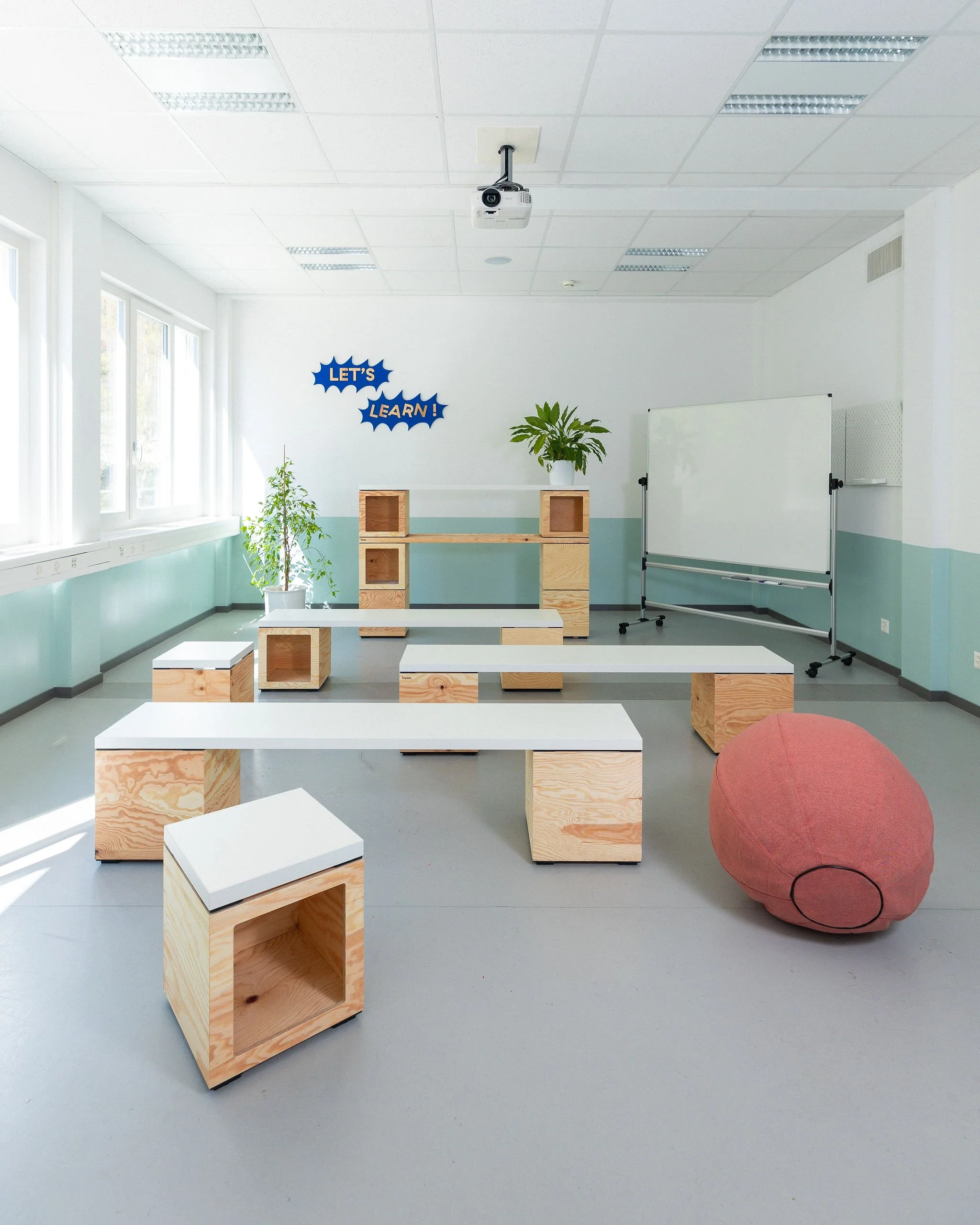 Une salle de workshop dans un coworking avec du mobilier modulable en bois clair, un bureau et étagères, des bancs, des plantes, un mur blanc avec une décoration en forme de nuages bleus avec des inscriptions en français « LET'S LEARN ! ».