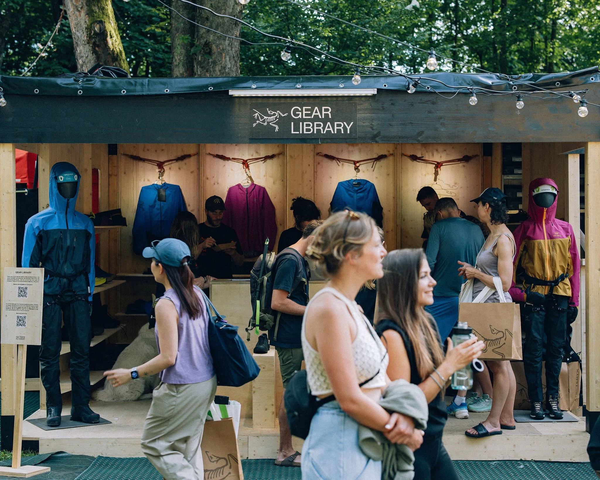Stand avec le signe 'Gear Library' en plein air, proposant des équipements de marche et des casques, avec plusieurs personnes intéressées autour.