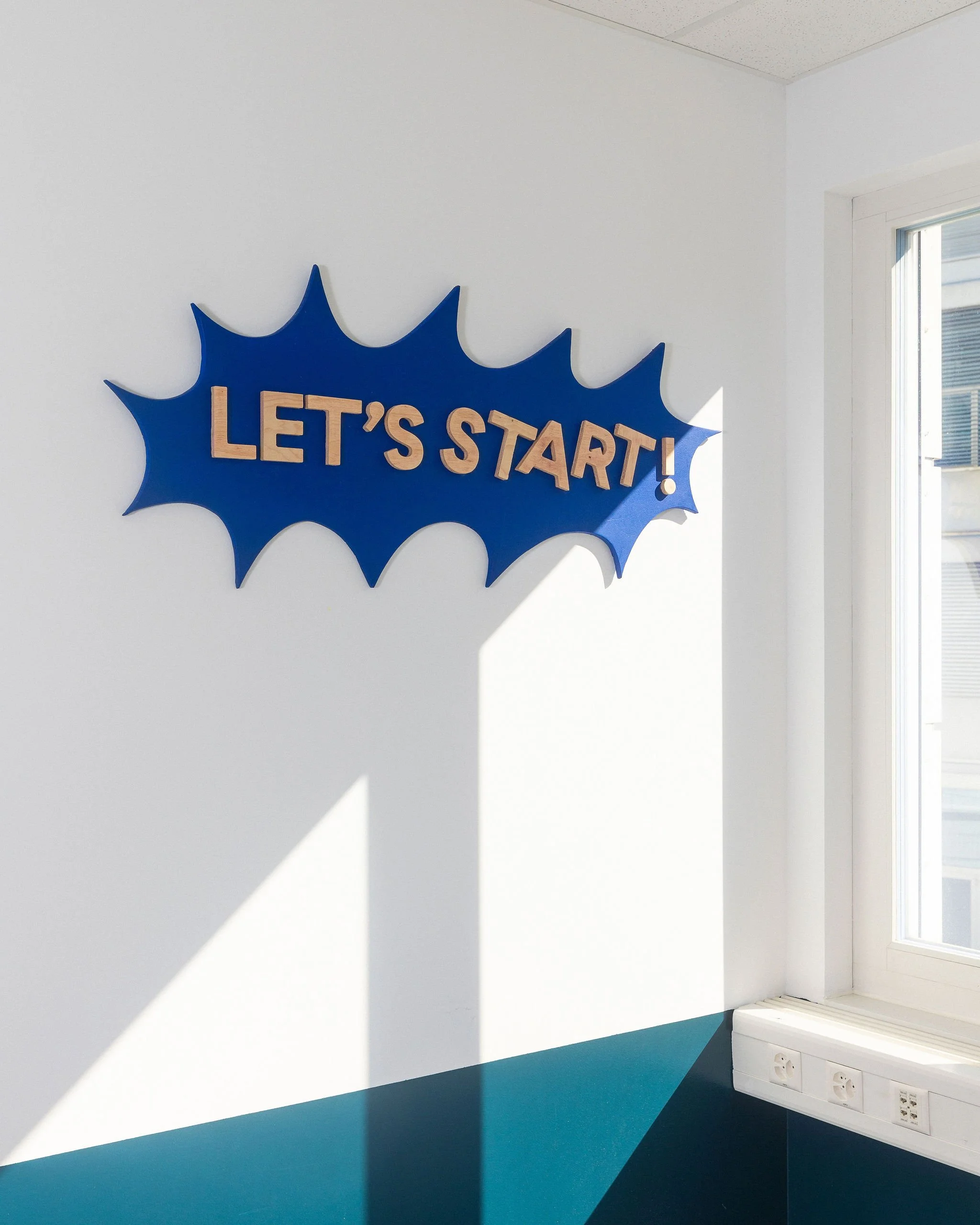 Mur blanc avec un panneau bleu en forme d'explosion et les mots 'LET'S START!' en lettres en bois. Fenêtre à droite, prises électriques en bas.