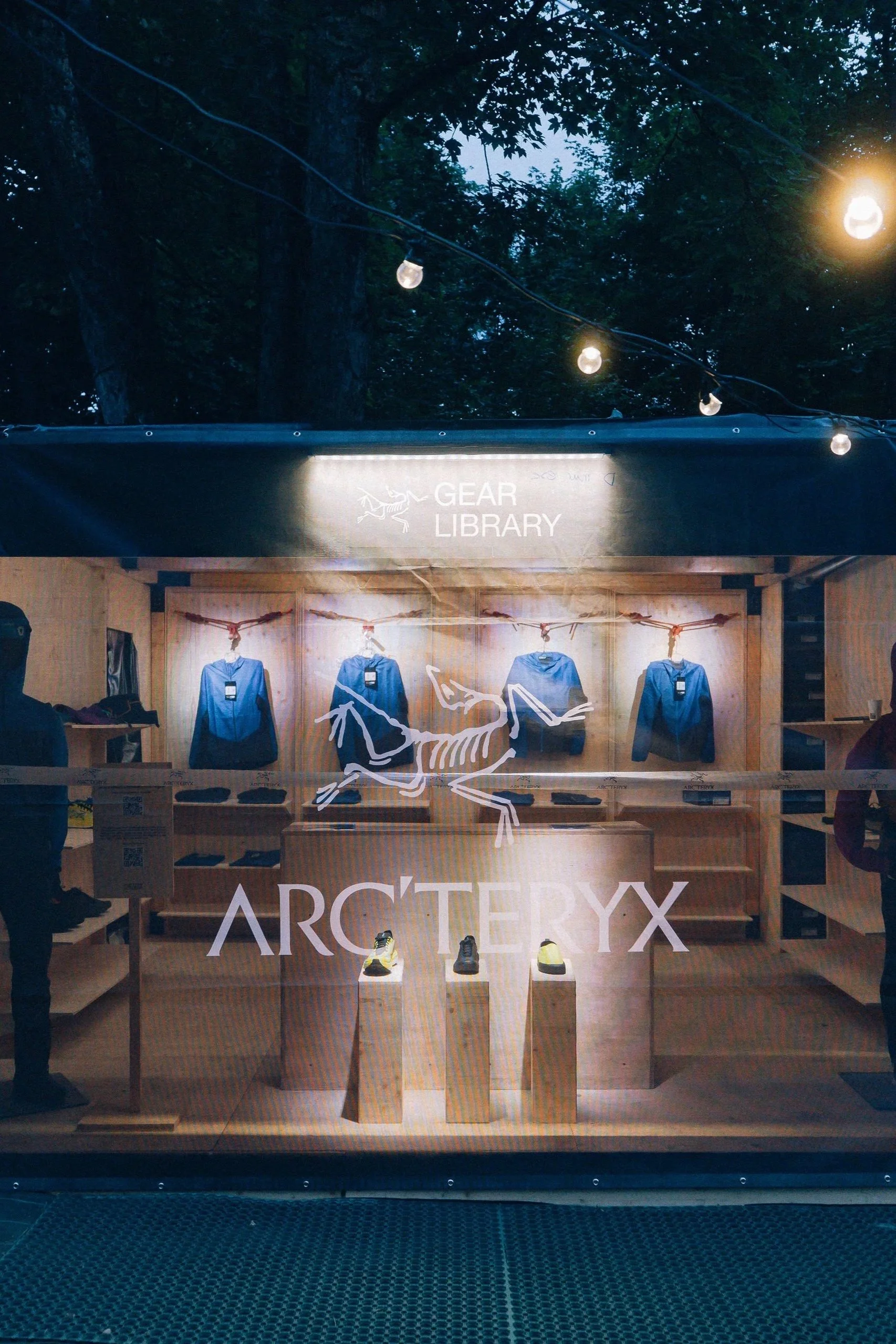 Vitrine d'une boutique Arc'teryx avec des vêtements de sport suspendus et des chaussures exposées en vitrine, éclairée par des lumières chaudes, dans un environnement de forêt.