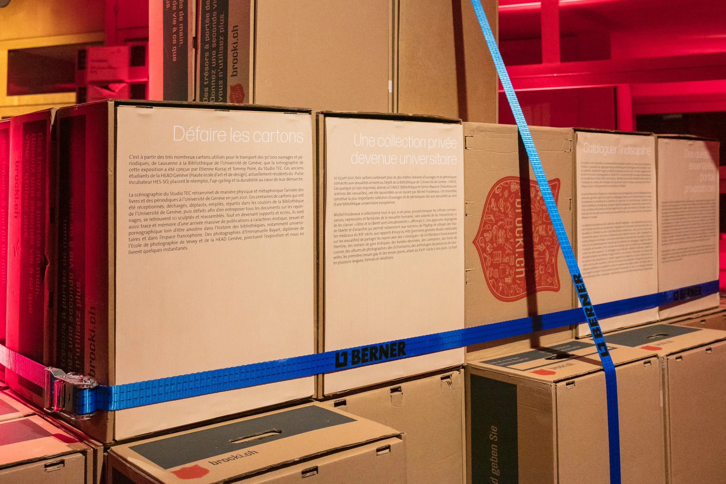 Une pile de cartons composent une scénographie dans une exposition sur la sexualité, emballés avec des sangles bleues. Sur les cartons on trouve le texte de présentation de l'exposition en français et des illustrations.