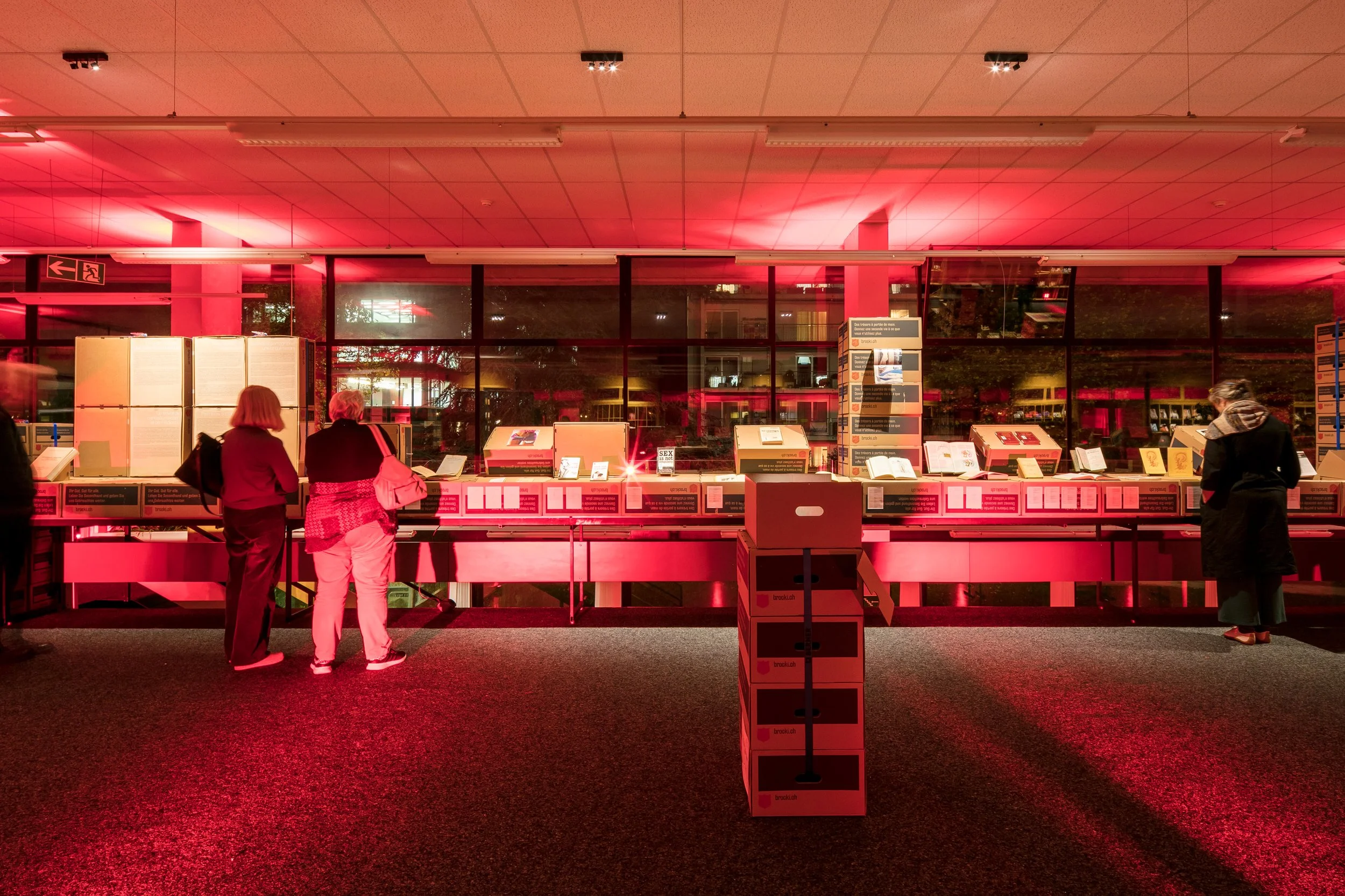 Salle d'accueil ou de vente avec des personnes regardant des livres ou des documents sur des tables, éclairée avec une lumière rouge, vue par une grande fenêtre la nuit.