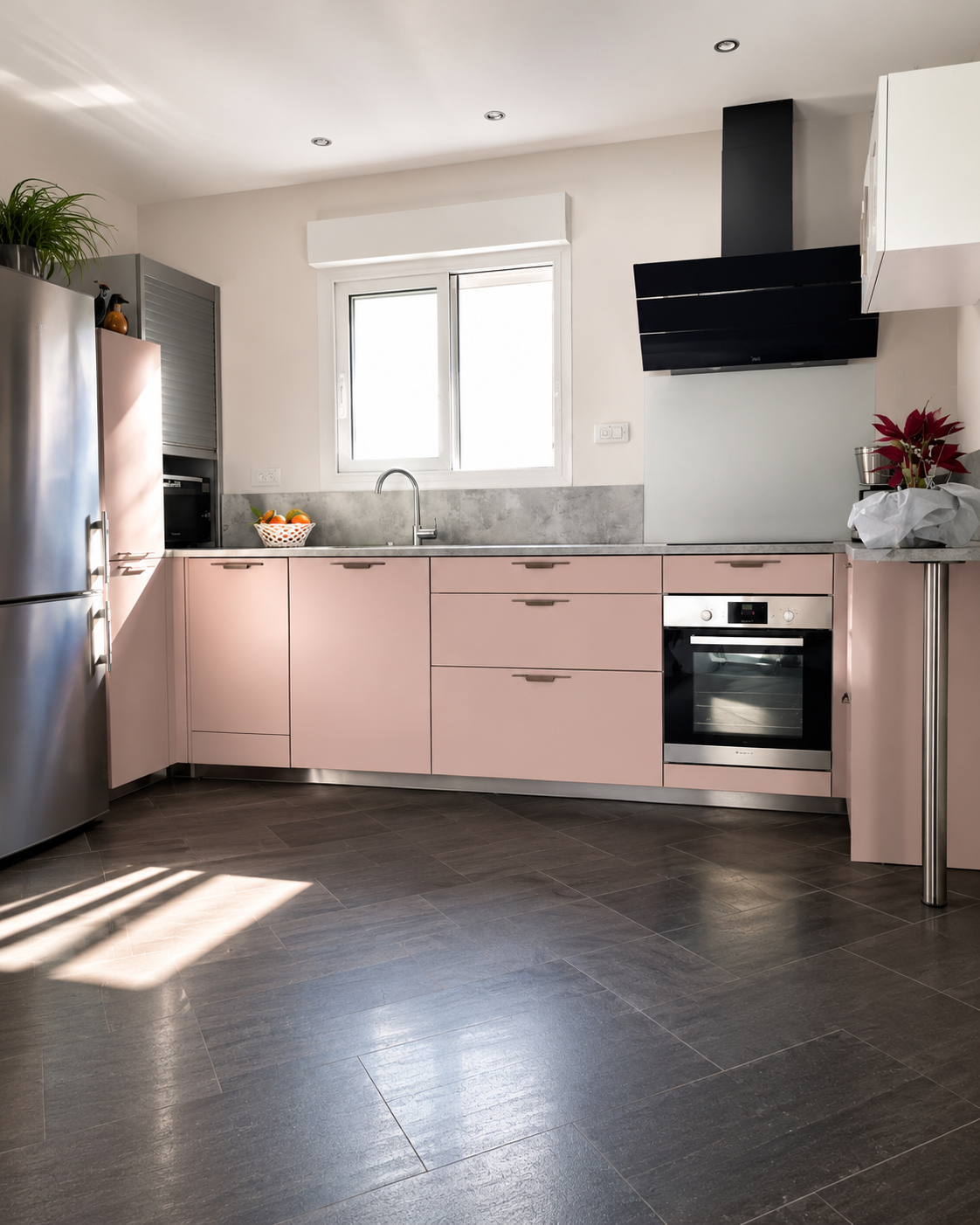 Cuisine moderne avec meubles roses, fenêtre, évier, four, hotte noire, réfrigérateur en acier inoxydable, plantes et fruits.