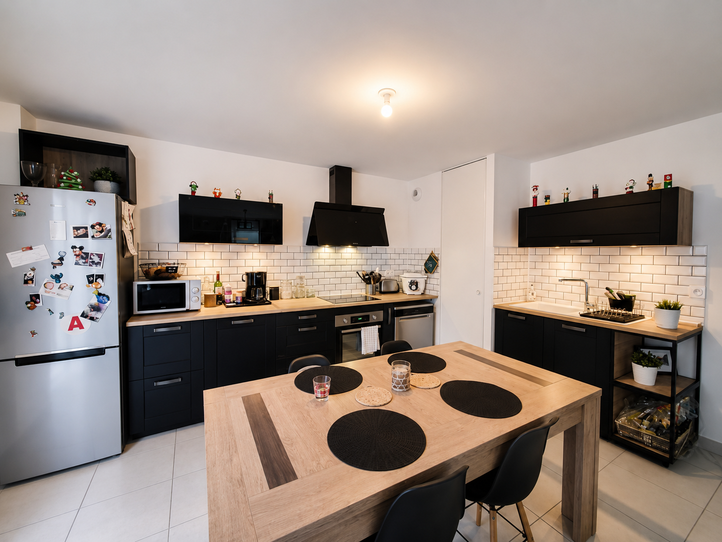 Cuisine moderne avec comptoirs noirs, meubles en bois clair, décor de Noël, table en bois avec quatre sets de table noirs, réfrigérateur en métal argenté avec aimants, appareils électroménagers, vaisselle, petits plants, et éléments de décoration.