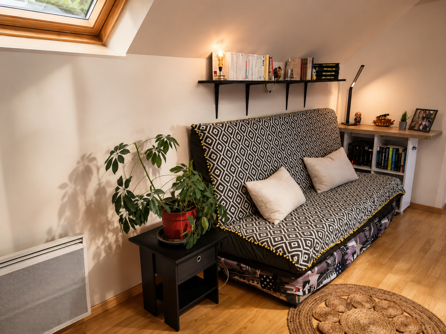 Salon avec un canapé recouvert d'une housse à motif géométrique noir et blanc, deux coussins beiges, une plante en pot rouge sur une petite table noire, un mur avec étagère à livres, une lampe et divers objets décoratifs, fenêtres de toit laissant en