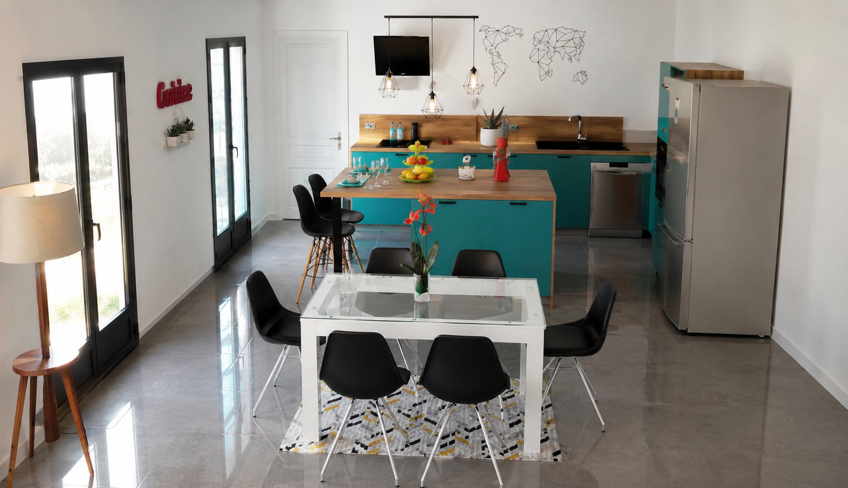 Une cuisine moderne avec un espace repas, comprenant une table blanche avec six chaises noires, une cuisine en bleu avec un îlot central en bois, un réfrigérateur en acier inoxydable, un meuble au mur, une télévision, et des décorations murales, dont