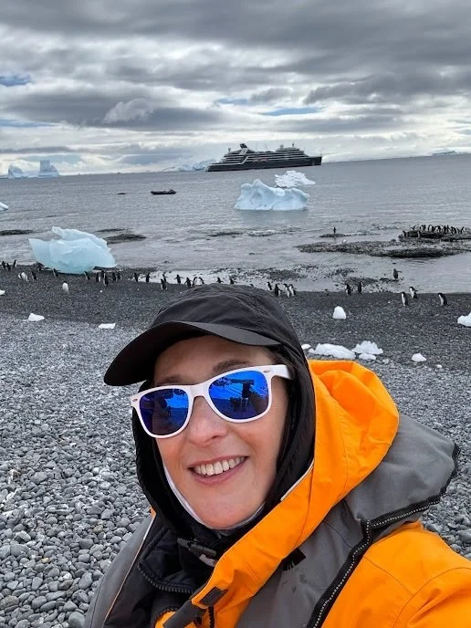Stephanie "PenguinsPreferred" Ng in an orange parka on shore in Antarctica, with penguins behind her