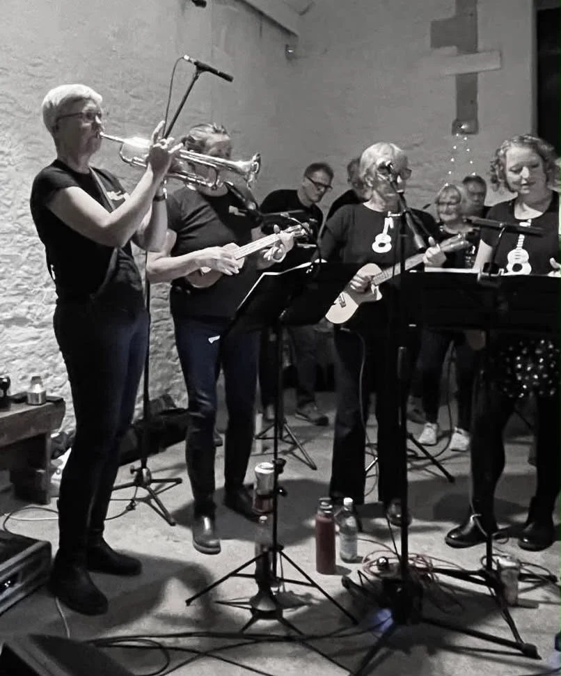 A group of people playing musical instruments, including a trumpet and guitars, in a band performance in a room with a stone wall.