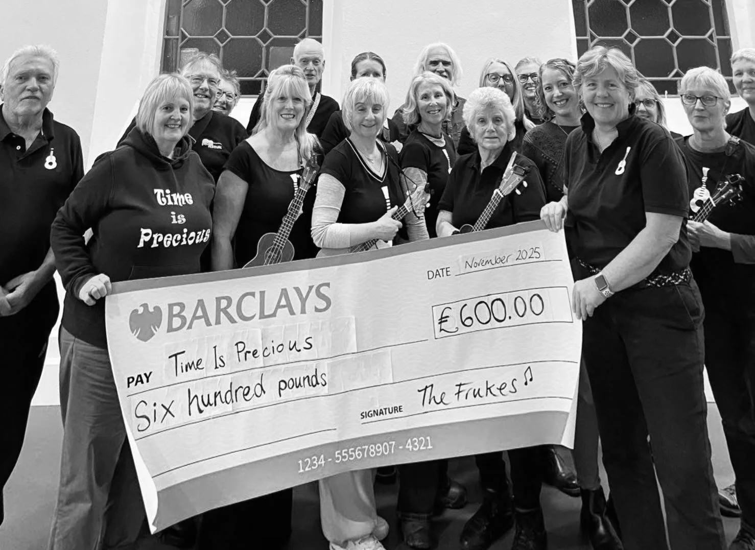 A group of people gathered indoors, holding a large check for 600 pounds from Barclays Bank, made out to 'Time is Precious' for the amount of six hundred pounds. The group includes both men and women, some holding ukuleles, and appears to be celebrating a donation or fundraising event.