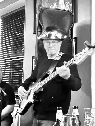 Older man playing electric guitar at an indoor gathering, with a large speaker and framed photos on the wall behind him.