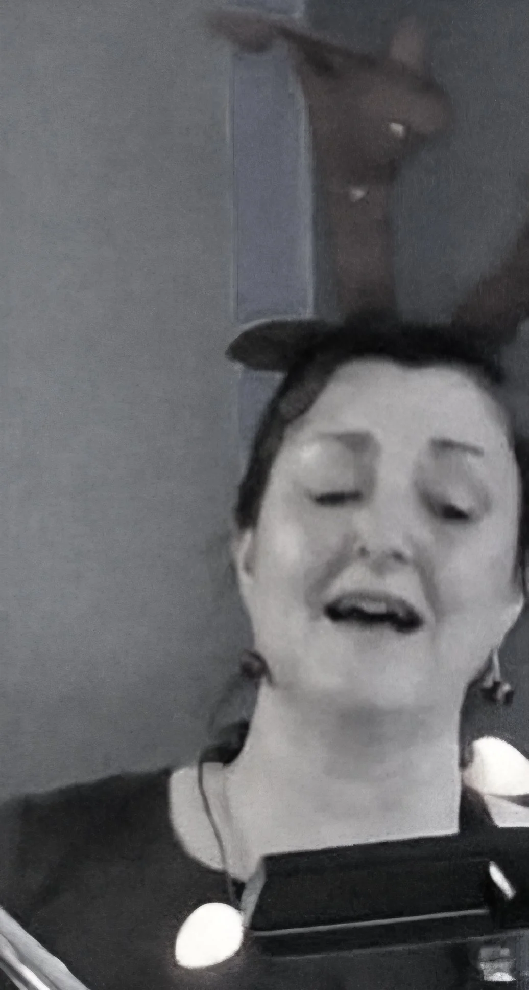 A black and white photo of a woman with her eyes closed, speaking or singing, with a large face mask in front of her resembling a person's face.