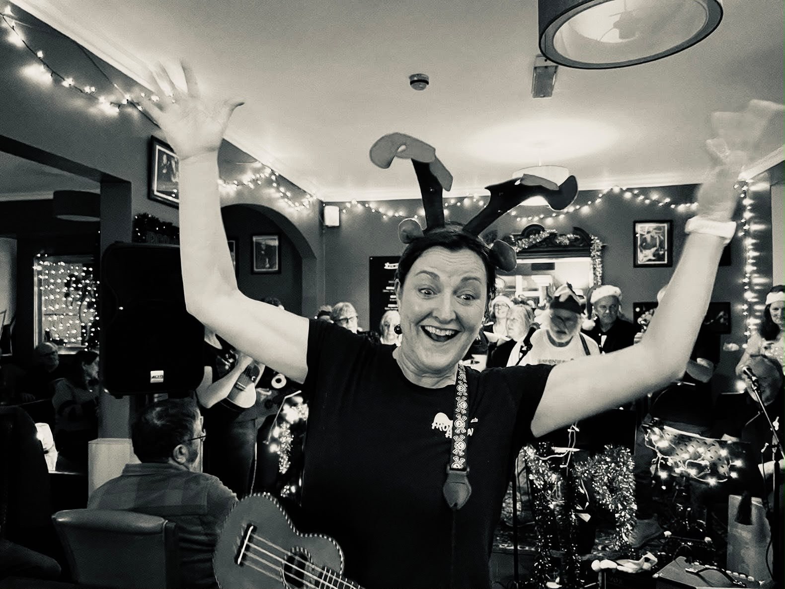 A woman celebrating at a holiday gathering, wearing reindeer antlers and a Christmas-themed necklace, with people in the background wearing Christmas hats and playing guitar, in a decorated room with string lights.