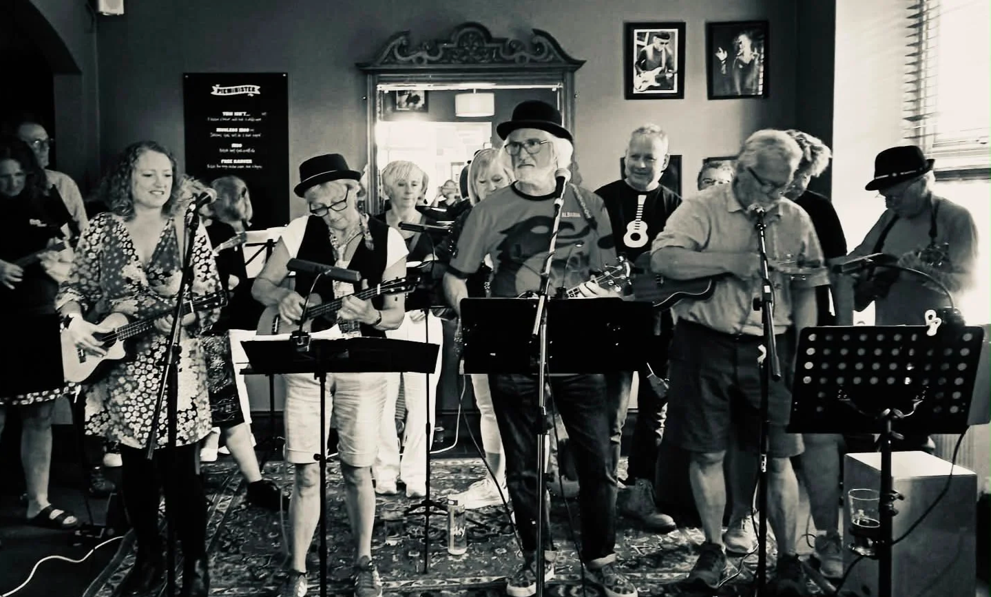 A group of musicians performing indoors, with some playing guitars and others singing, in a room decorated with photographs and artwork on the walls.