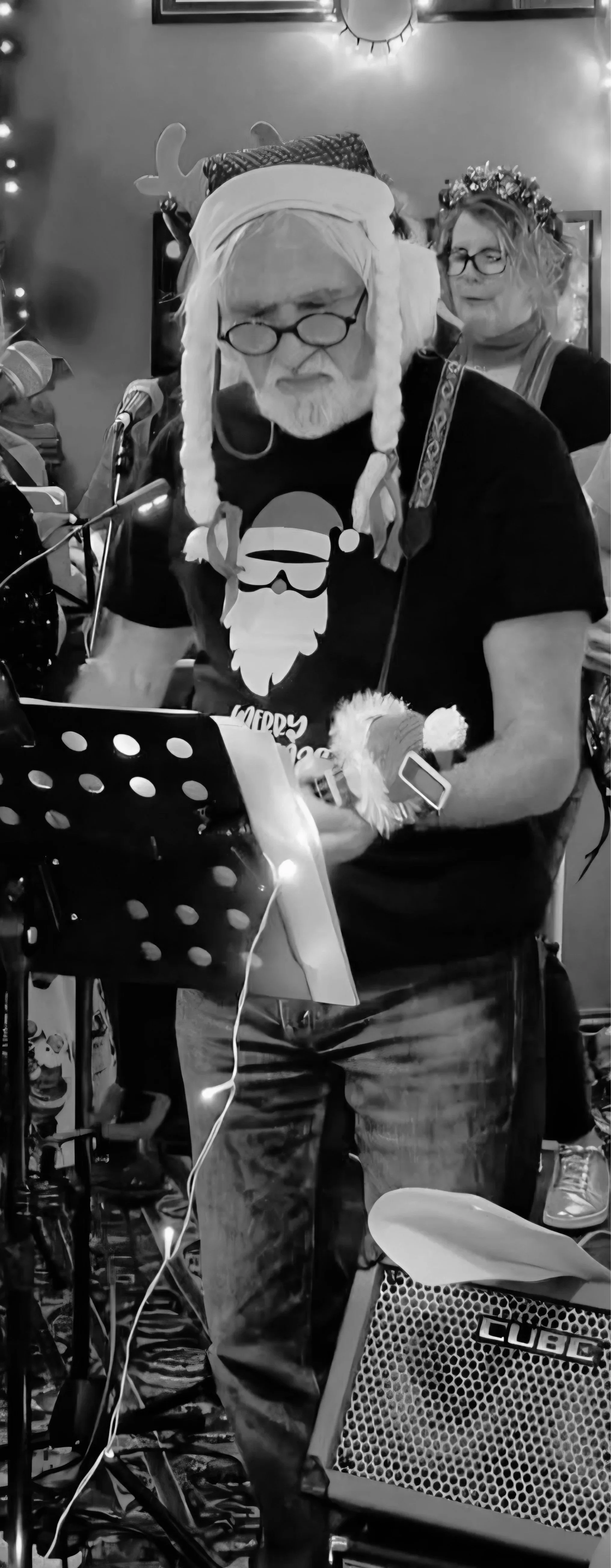 A man dressed in Christmas-themed attire with a Santa hat, reindeer antlers, and holiday glasses, playing music with a microphone and music stand, surrounded by festive decorations and on a stage.