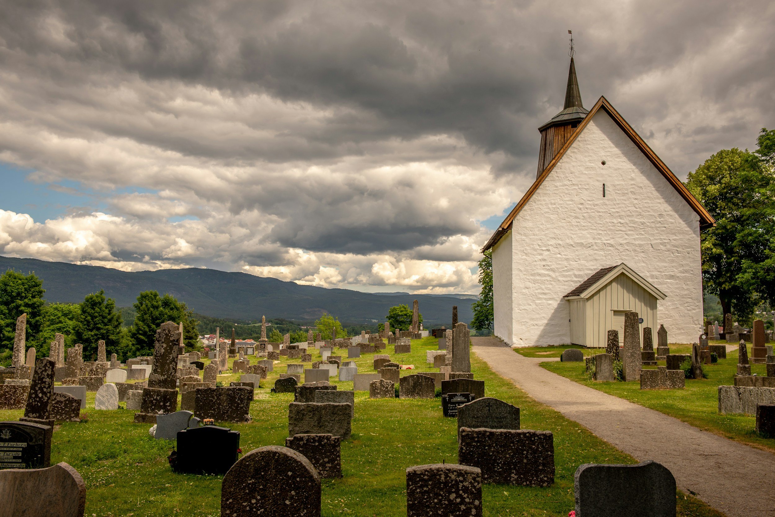 A white church with a pointed steeple surrounded by a graveyard with numerous headstones under a cloudy sky.