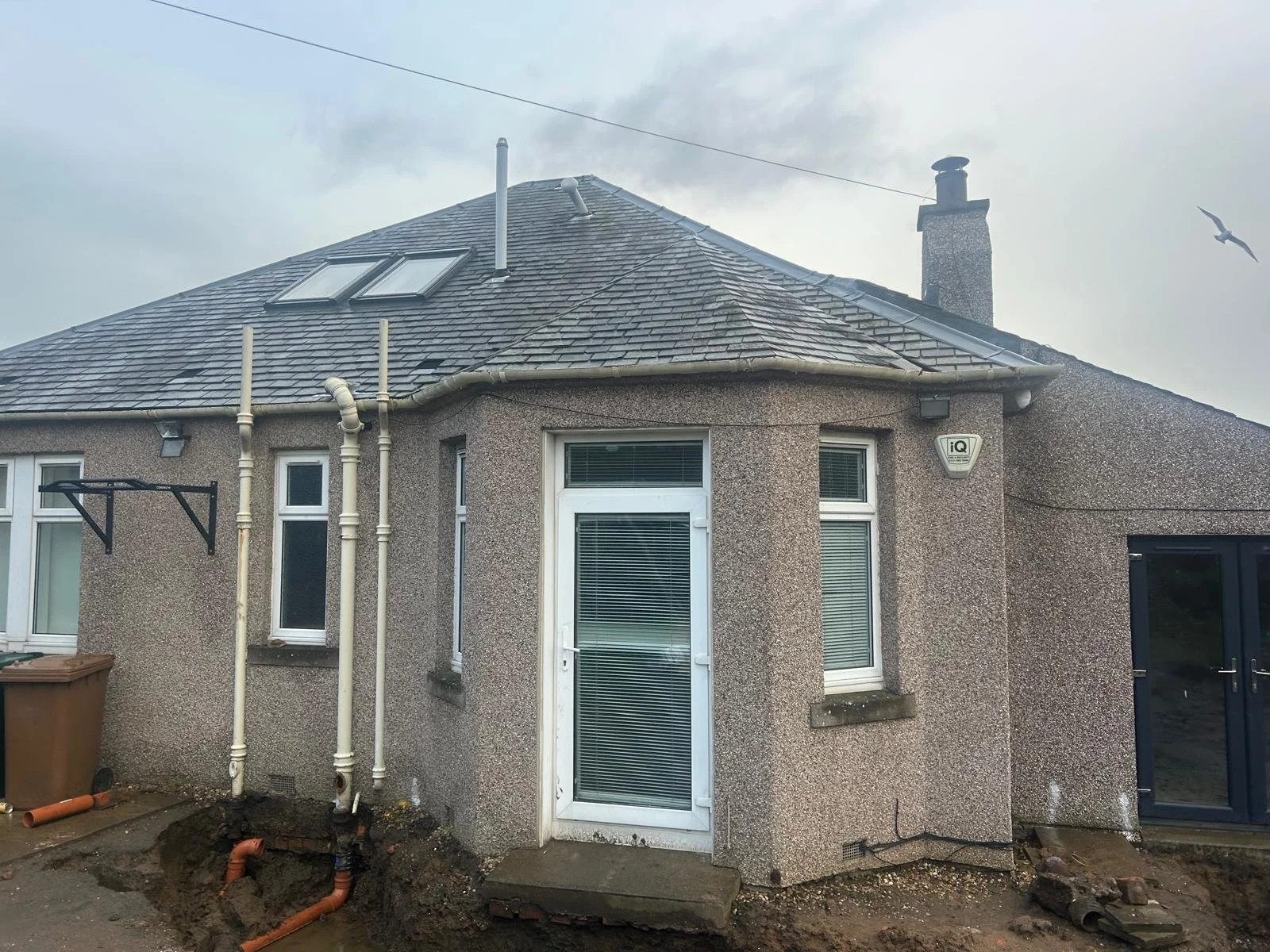 Back of a house with a tiled roof, two skylights, and white-framed windows. Some pipes are visible on the wall, and the ground appears under construction with dirt and pipes.