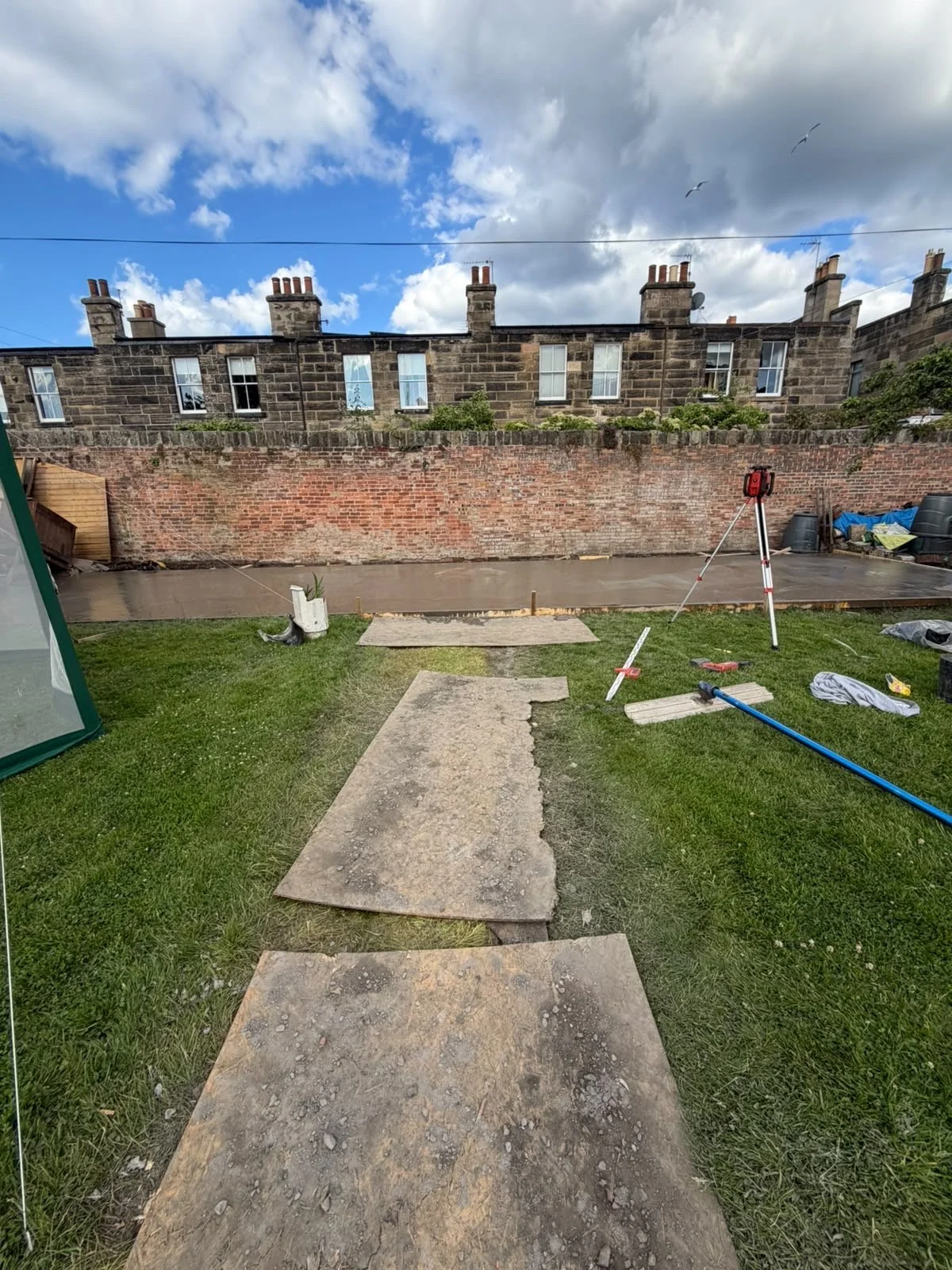 A backyard with a gravel walkway leading to a cement patio, construction tools and materials, and a brick wall with houses in the background under a partly cloudy sky.