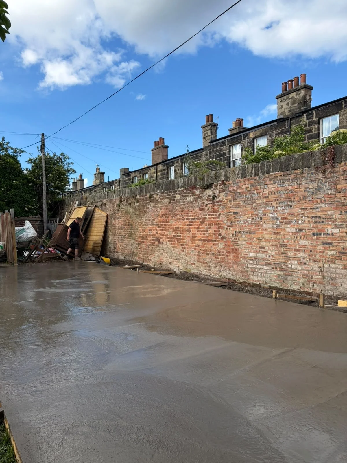 A person working on a newly poured concrete surface in a backyard with a brick wall and townhouse buildings with chimneys in the background.