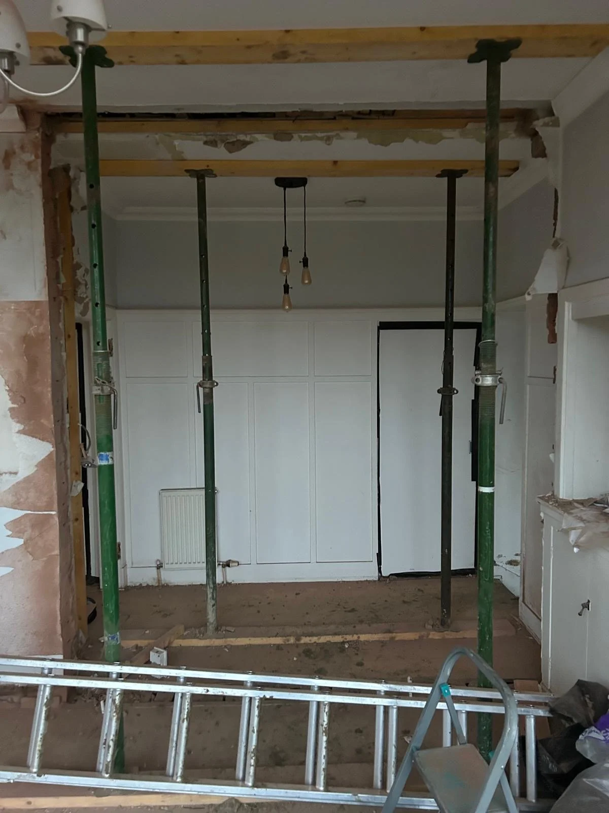Room under renovation with exposed wall and ceiling framing, construction support poles, a ladder, and incomplete wall coverings.