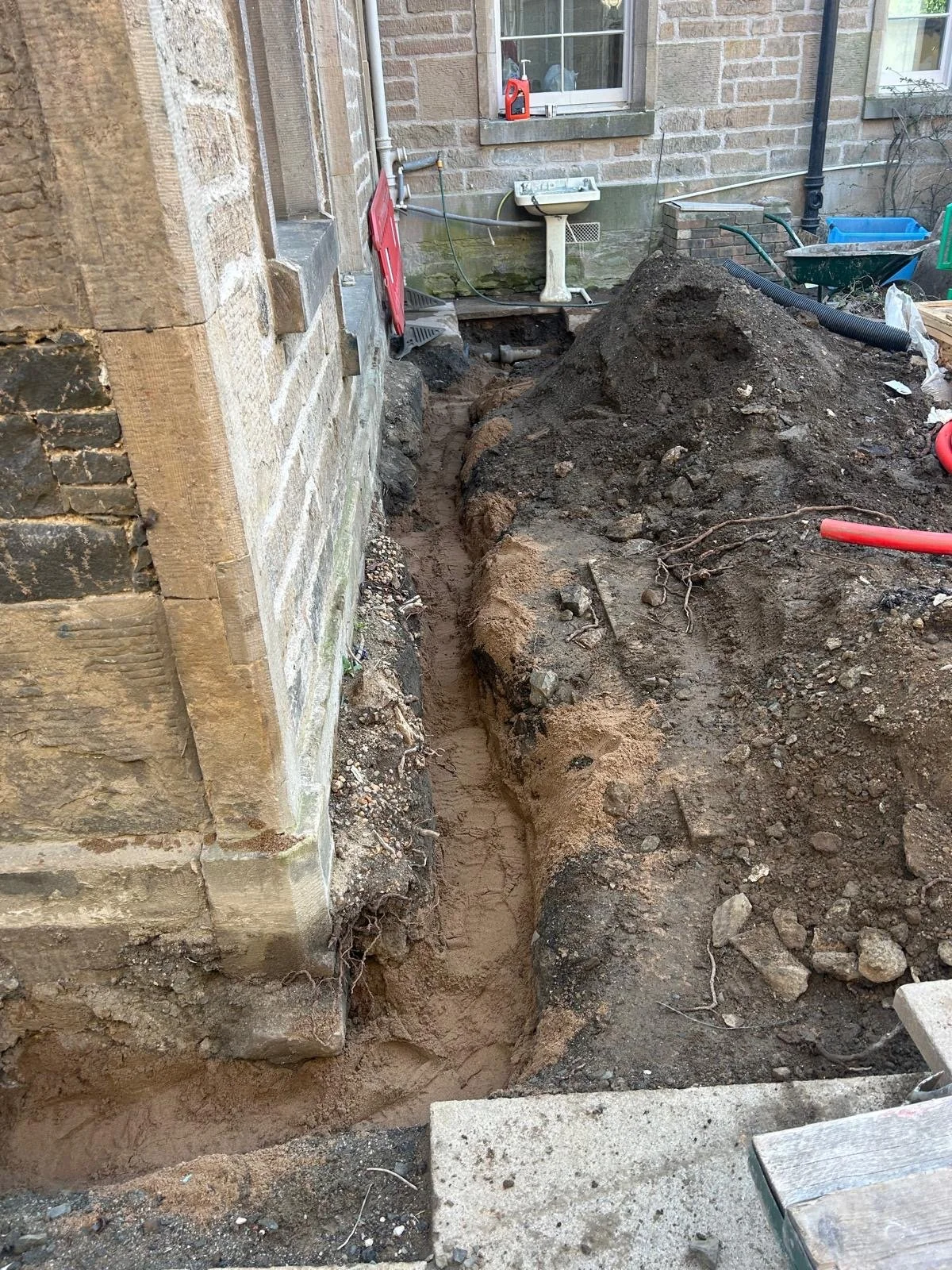 A construction or excavation site outside a brick house with loose soil and dirt, showing a trench dug along the side of the house's foundation.