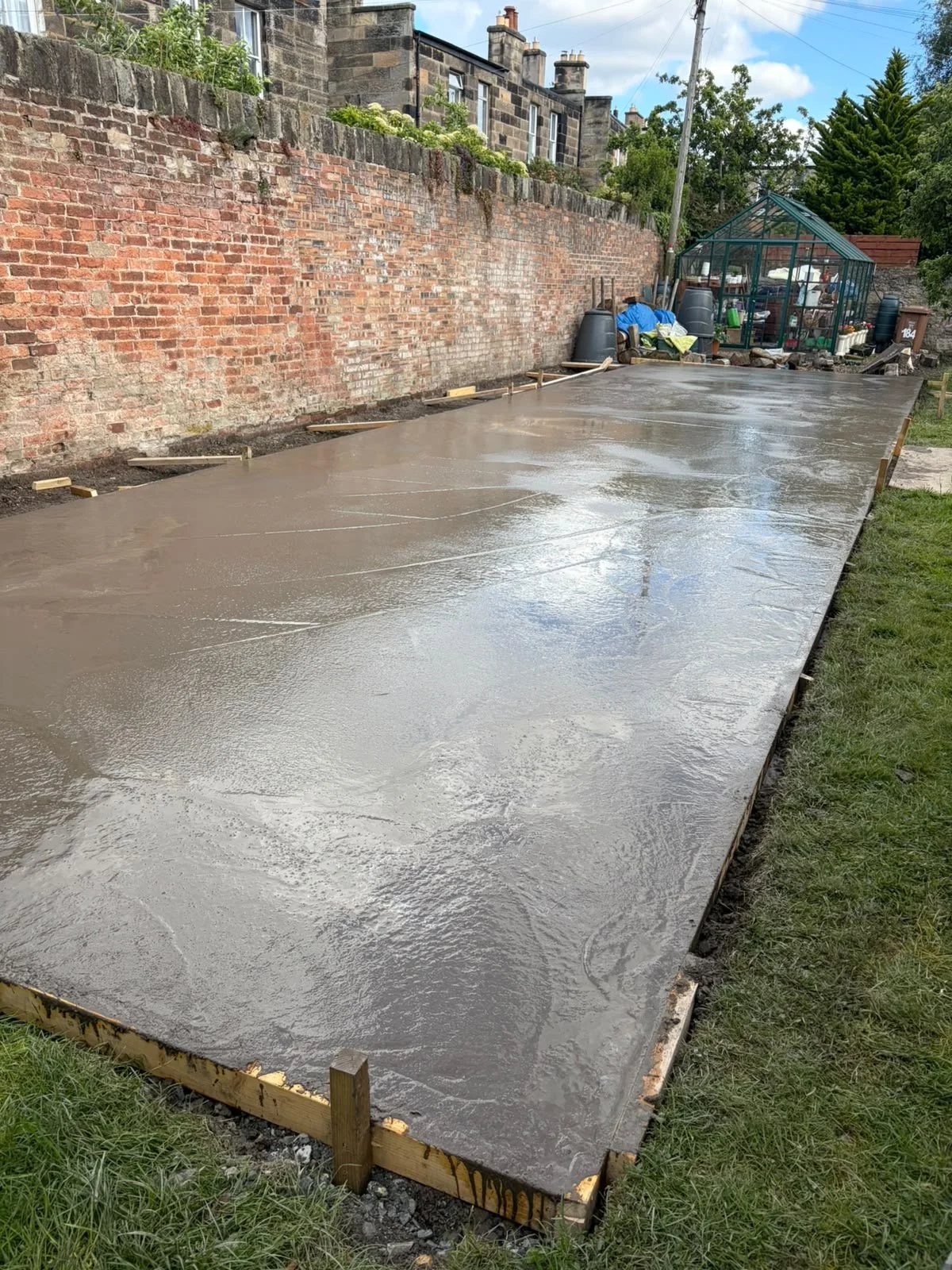 Freshly poured concrete slab in a backyard with a brick wall and a greenhouse in the background.