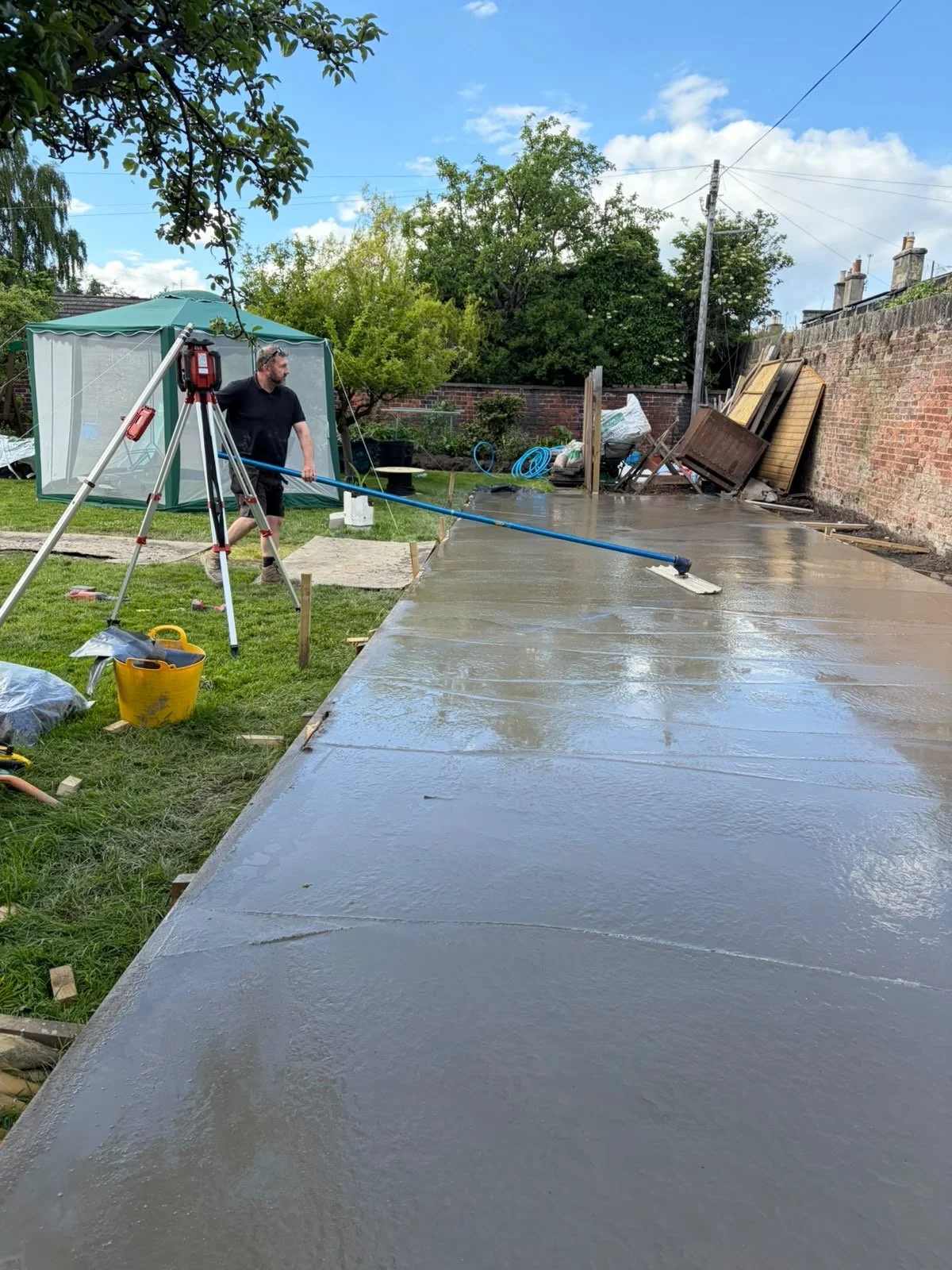 A man is using a power washer to clean a recently poured concrete patio in a backyard, with construction materials and tools nearby.