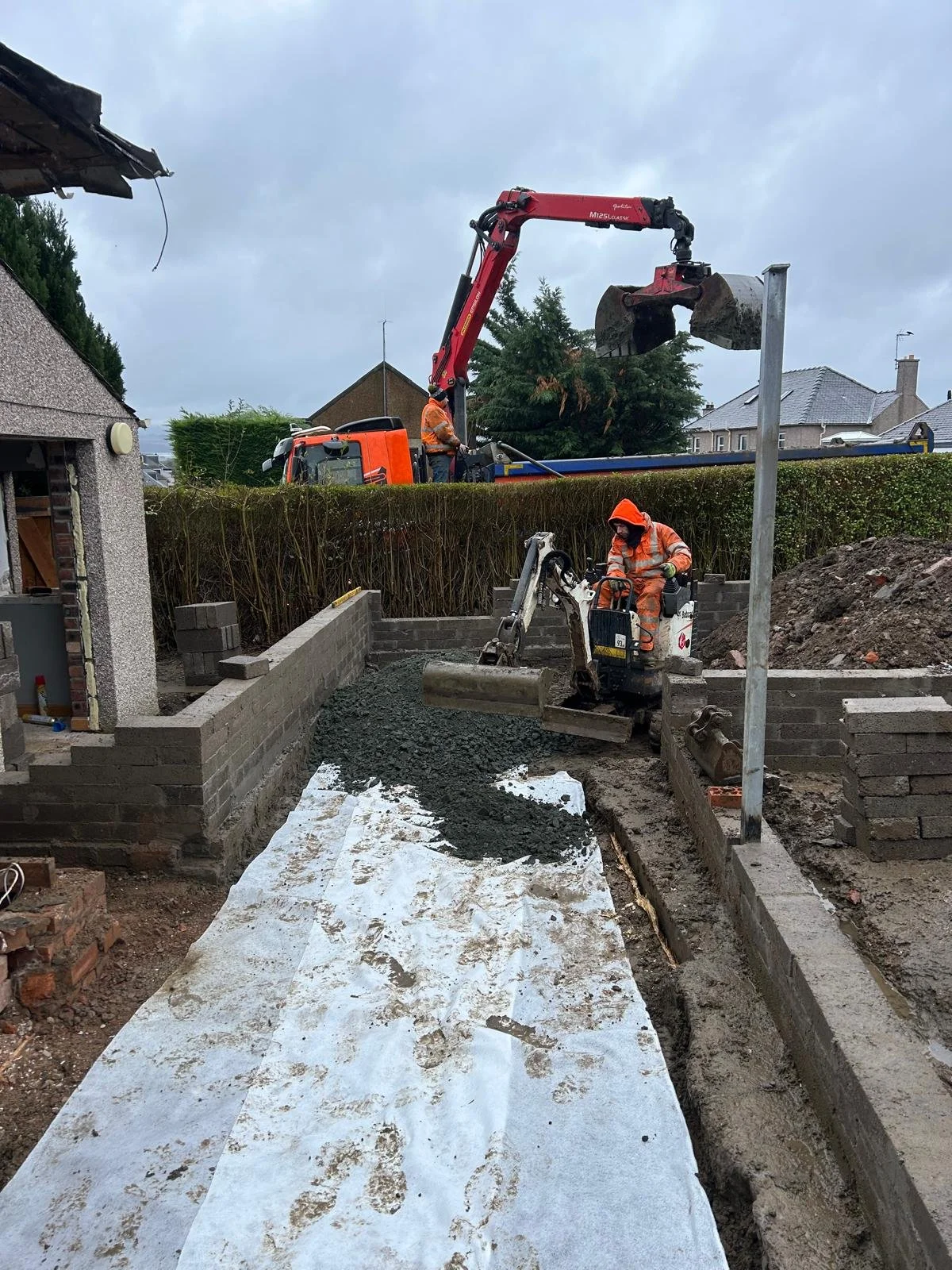 Construction workers in orange safety gear working on a building foundation. One worker is operating a small excavator, while another is using a machine to pour concrete onto a prepared surface. There is an excavator with a long arm and bucket in the background, with a truck nearby unloading gravel or concrete. The site is partially enclosed by a brick wall, and there are residential buildings and trees in the background under a cloudy sky.