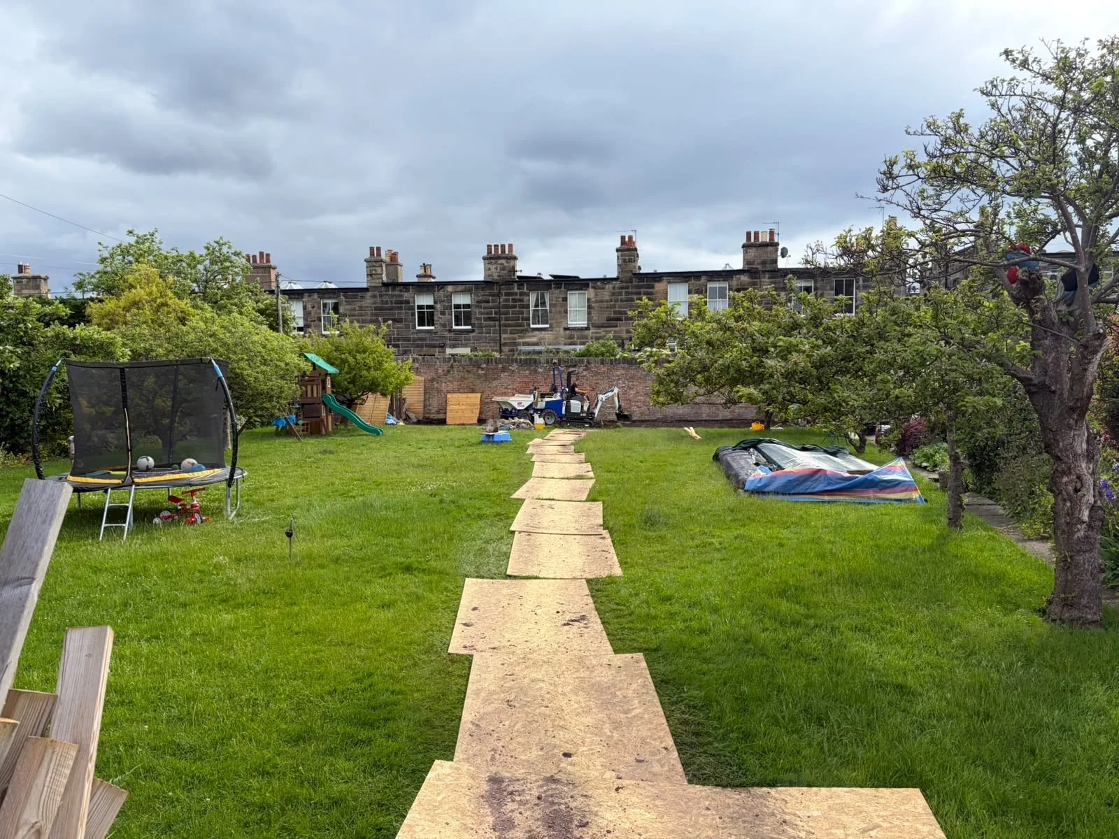 A backyard with a narrow stone pathway leading to a brick wall, surrounded by green grass, trees, a trampoline, and outdoor toys, under cloudy sky.