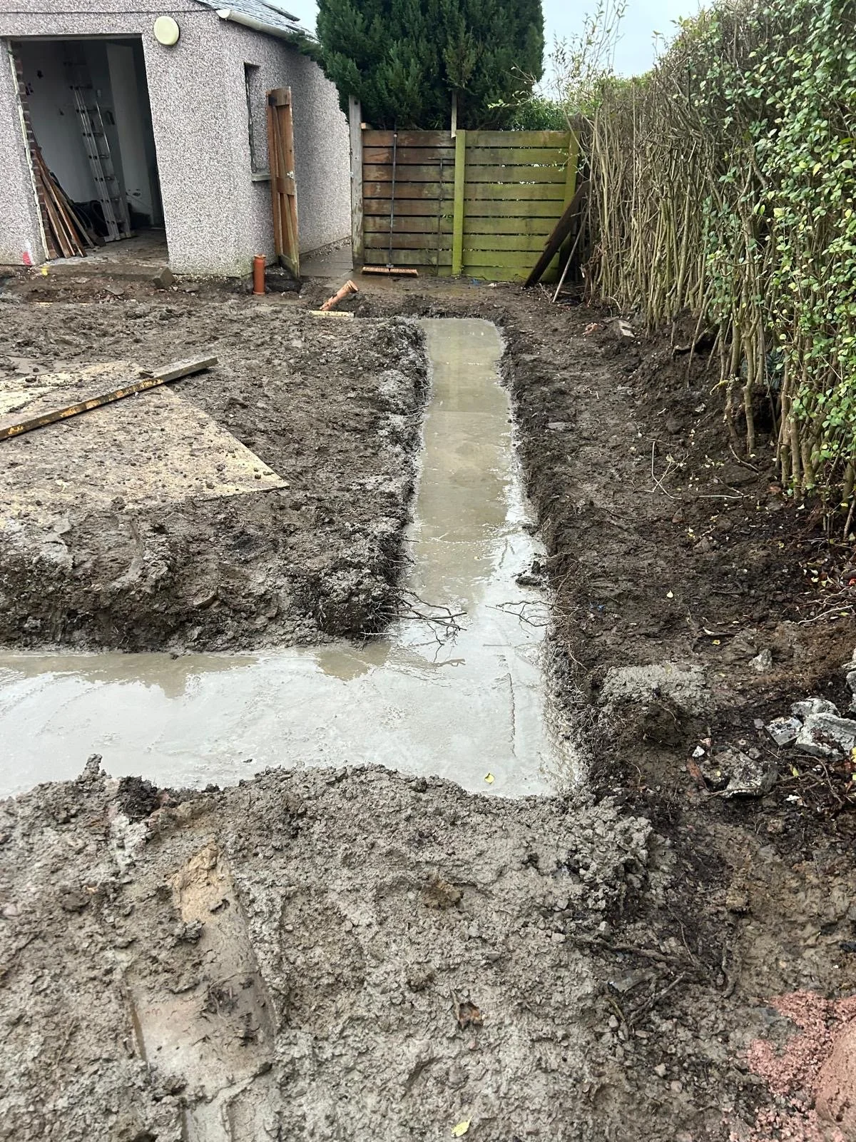 A backyard area under construction with freshly poured concrete for a pathway, surrounded by excavated dirt and a wooden fence on one side and bushes on the other.