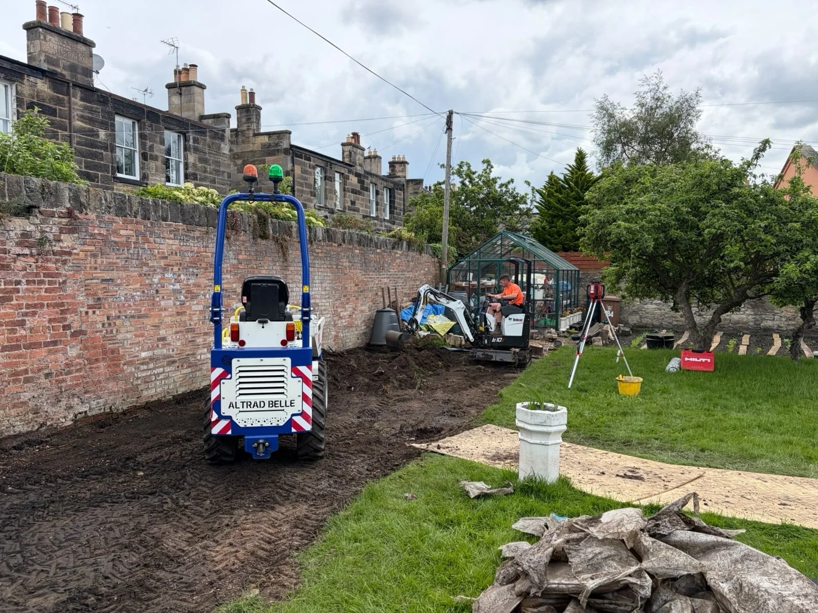 A backyard garden under construction with a small excavator, a person operating it, a green greenhouse, a brick wall, and construction tools and materials, including a wheelbarrow, buckets, and large rocks.