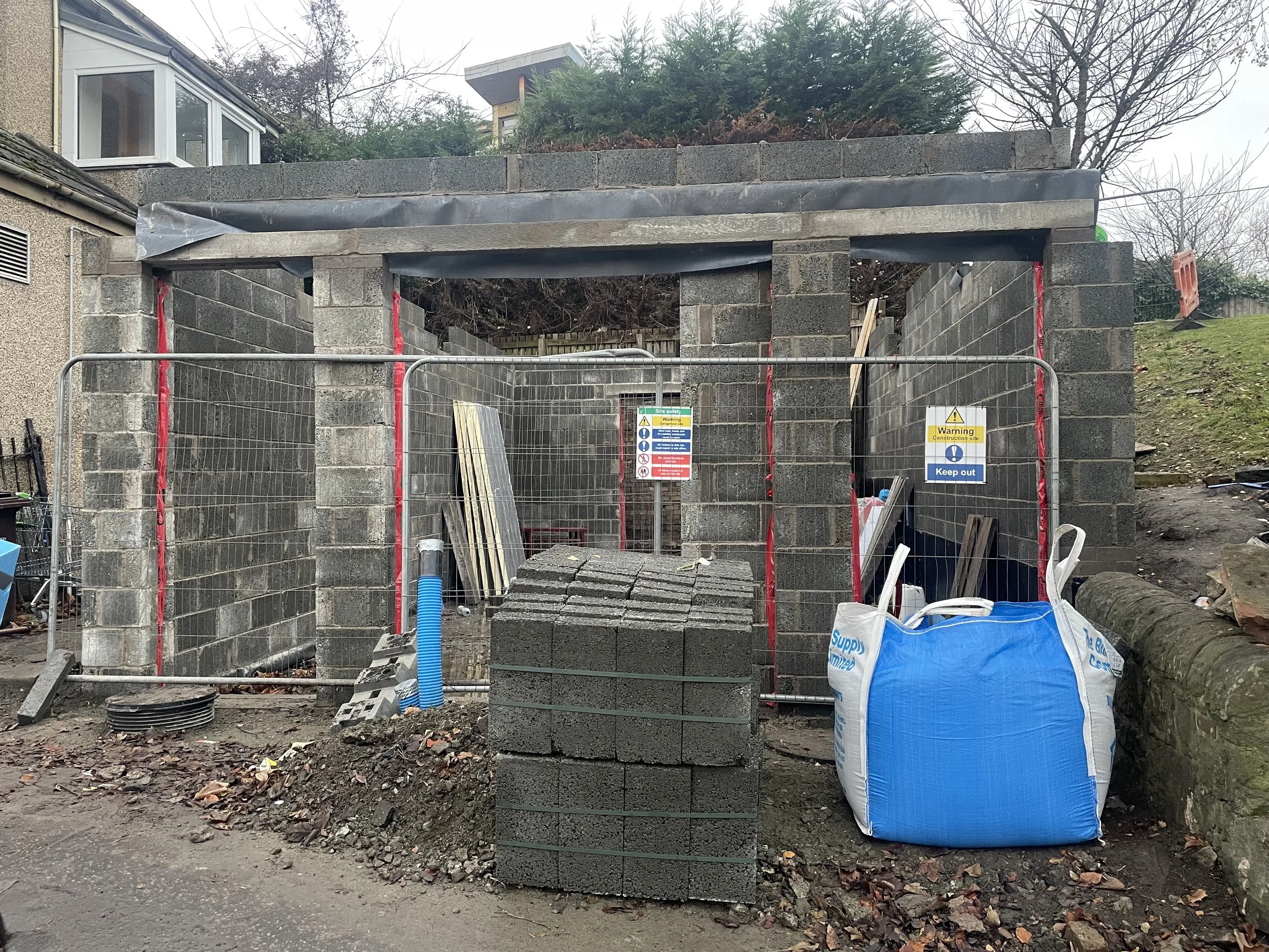 Under construction site with grey cinder block walls, construction fencing, and construction signs. Building materials and equipment are present, and the area is surrounded by trees and nearby houses.