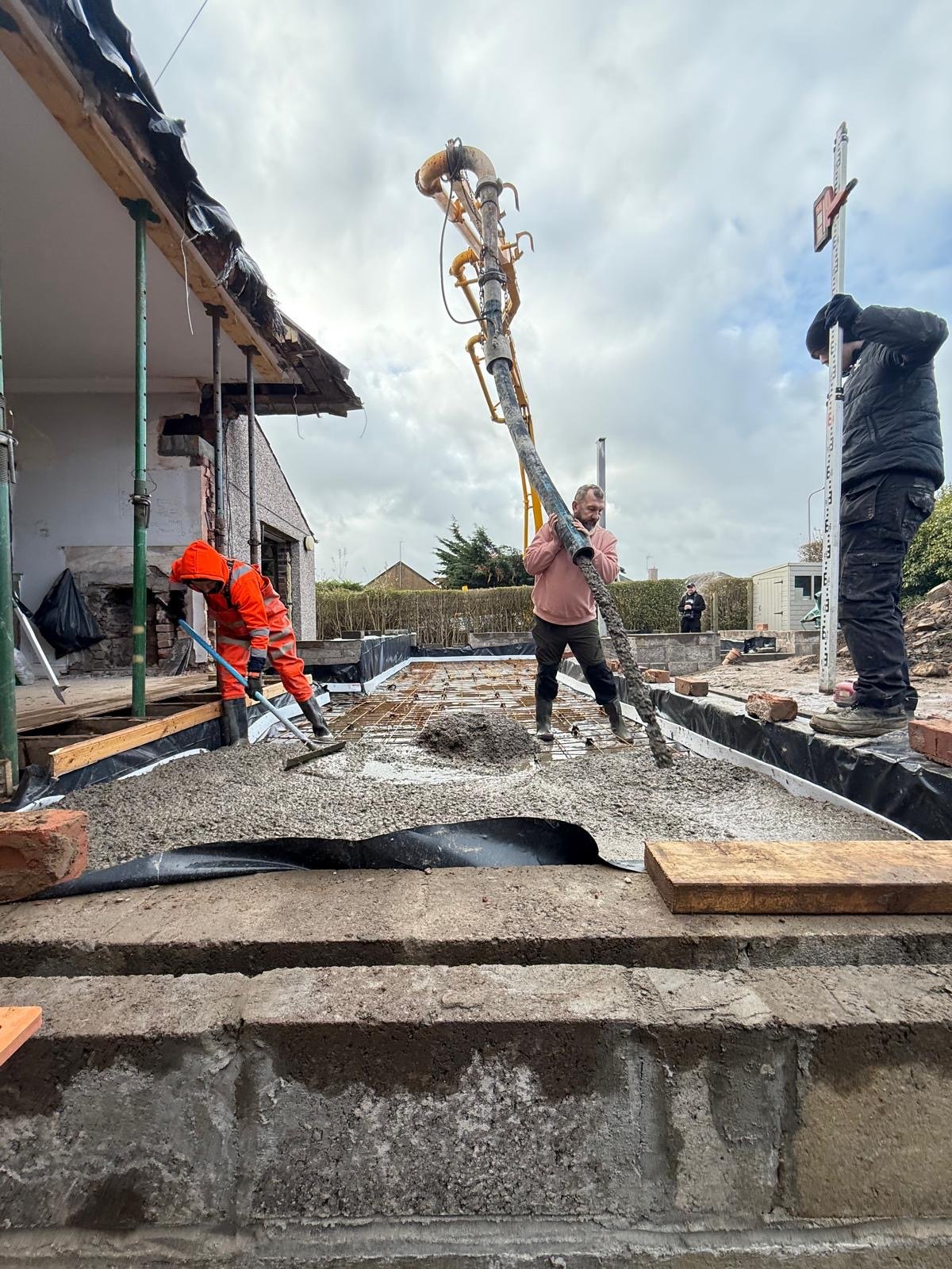 Construction workers pouring and leveling concrete on a building site foundation, with a concrete pump in operation.