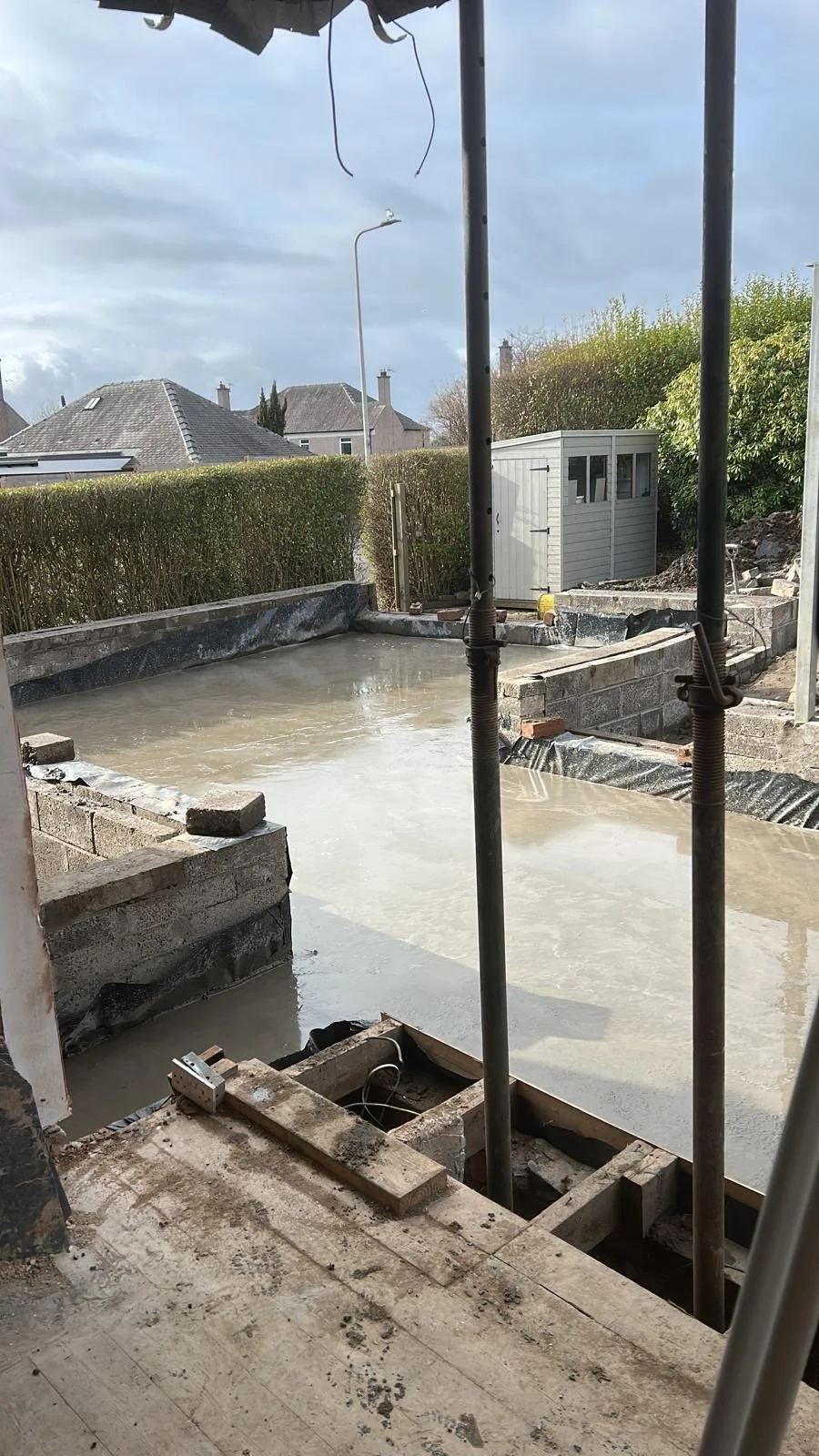 A construction site with freshly poured concrete for a patio or foundation, with building materials and scaffolding in place, and a background of residential houses and a hedge.