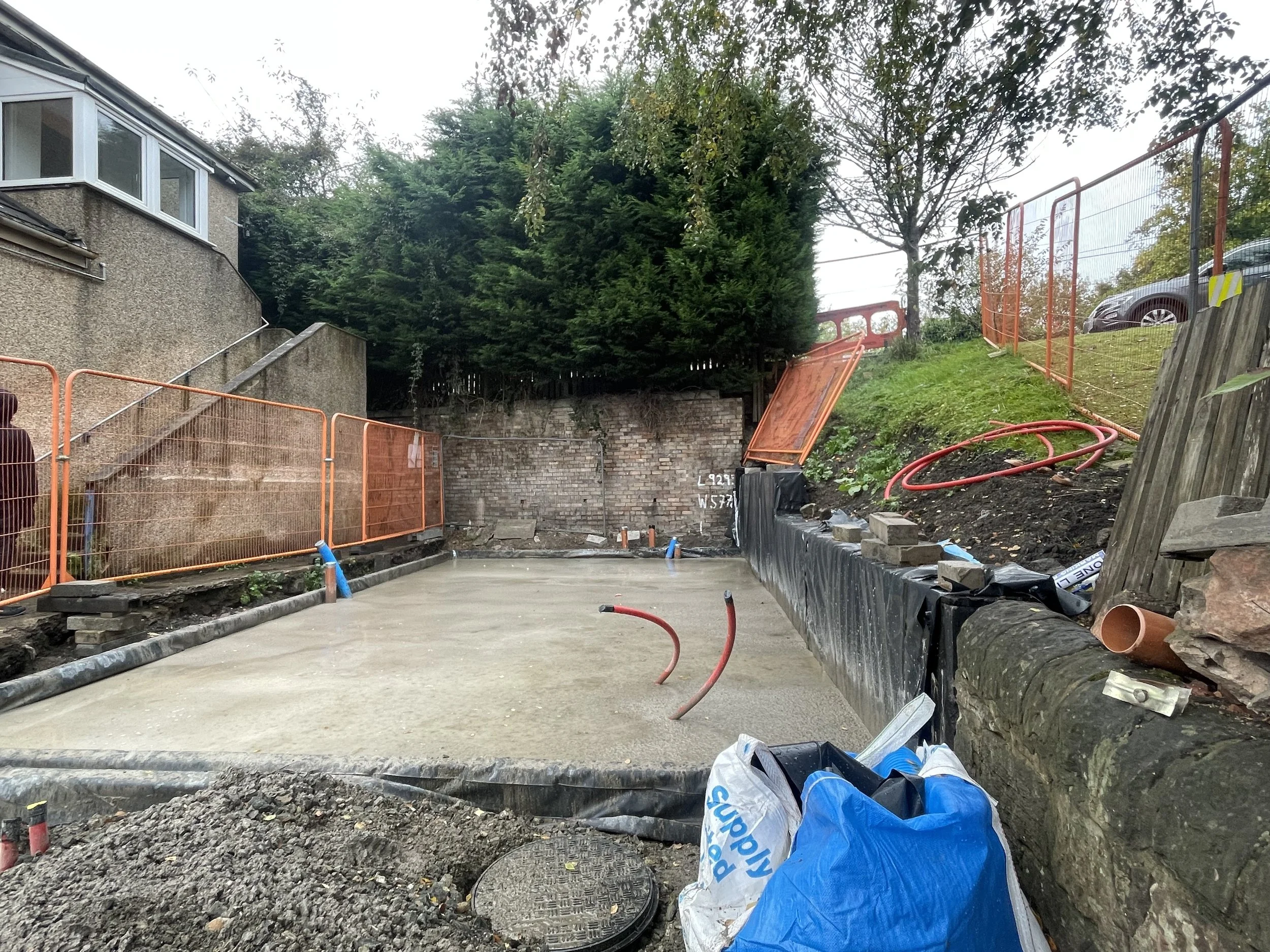 A construction site with freshly poured concrete, orange safety fencing, and construction materials such as hoses and bags in the foreground. There are trees and a house on the left, with a brick wall in the background.
