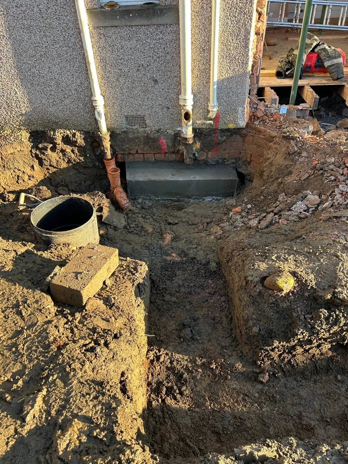 Construction site with dug trench, exposed pipes, and bricks, possibly for plumbing or foundation work.