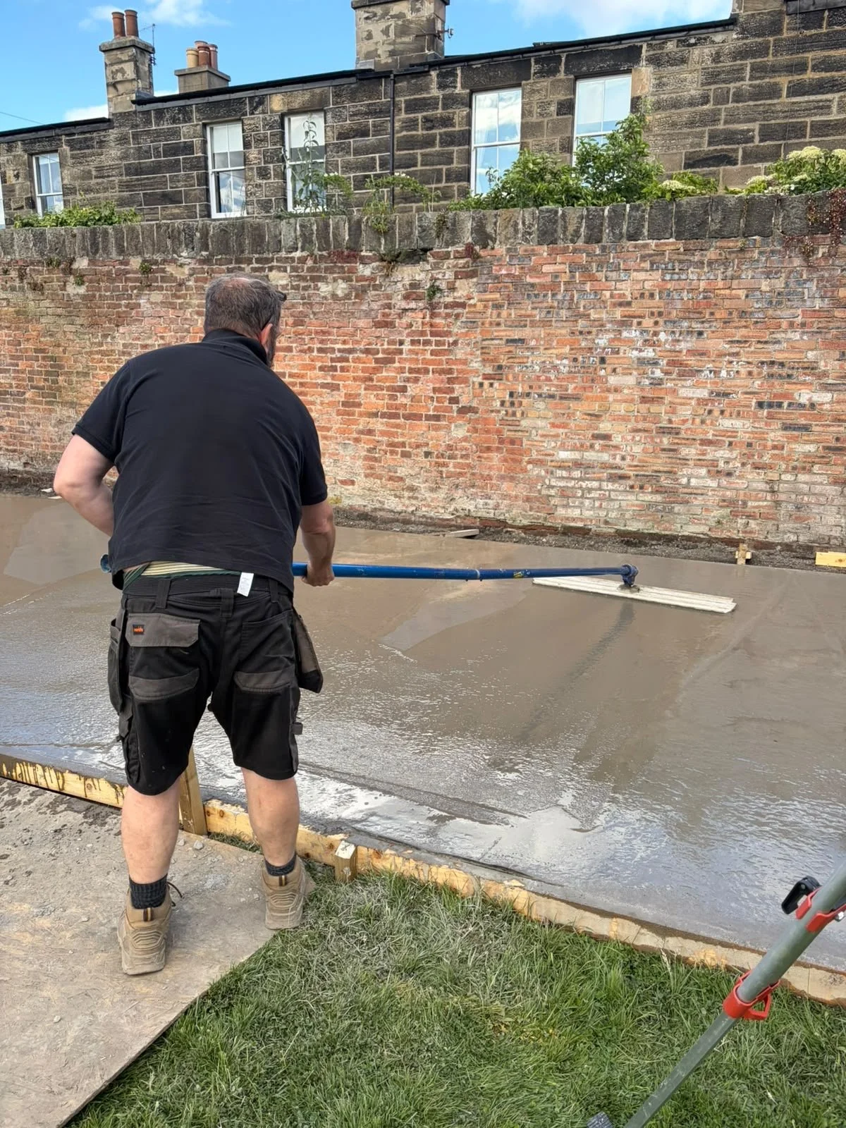 A man wearing a black shirt and black shorts is using a long hand tool to level freshly poured concrete on a construction site outdoors. There is a brick wall, a stone building with windows, and a brick wall with plants behind the man.
