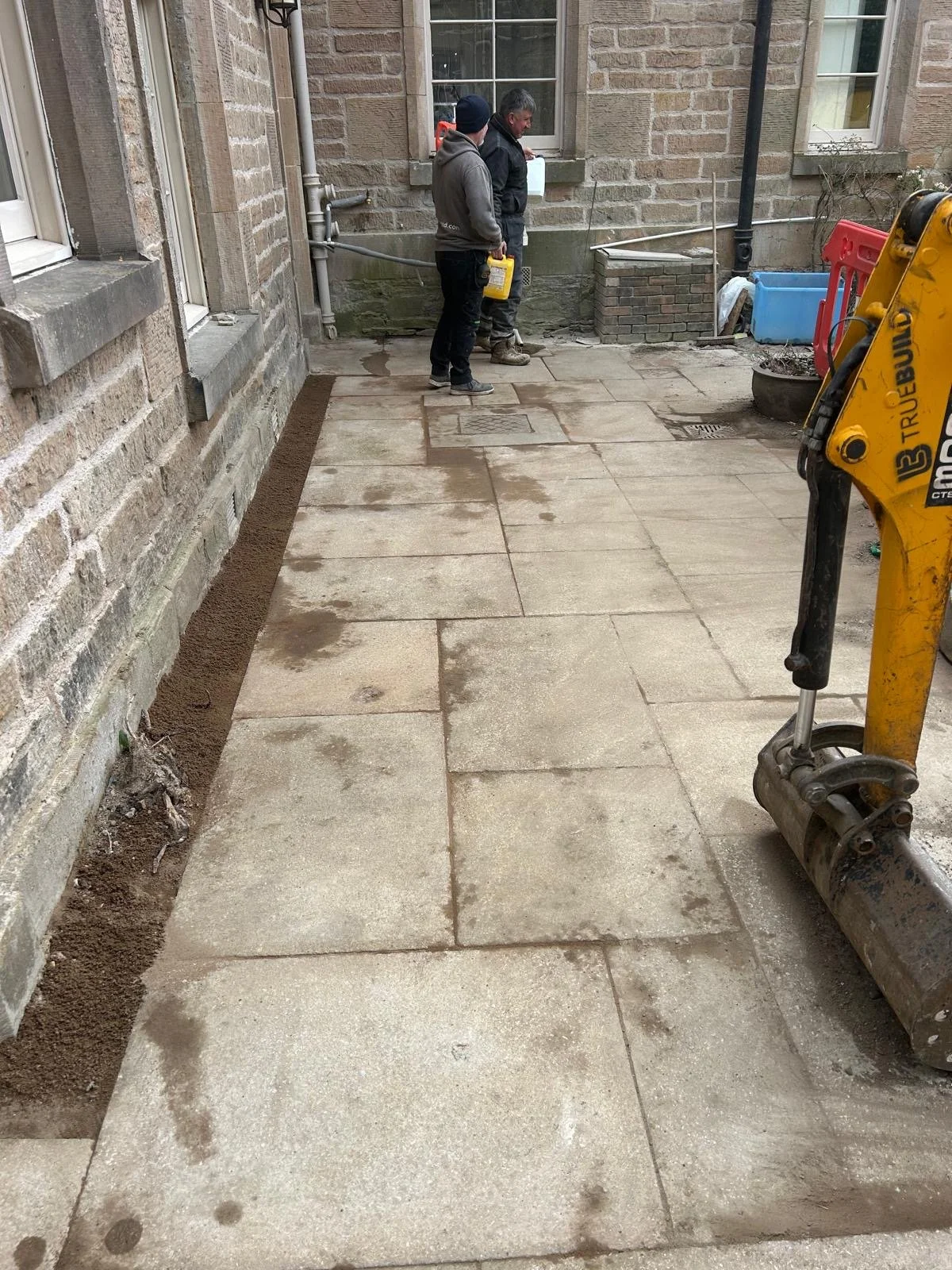Two workers are installing a new stone patio, with one holding a container and the other holding a spraying bottle, next to an older brick building. A small excavator is in the foreground, and the area along the edge is being prepared for laying stones.