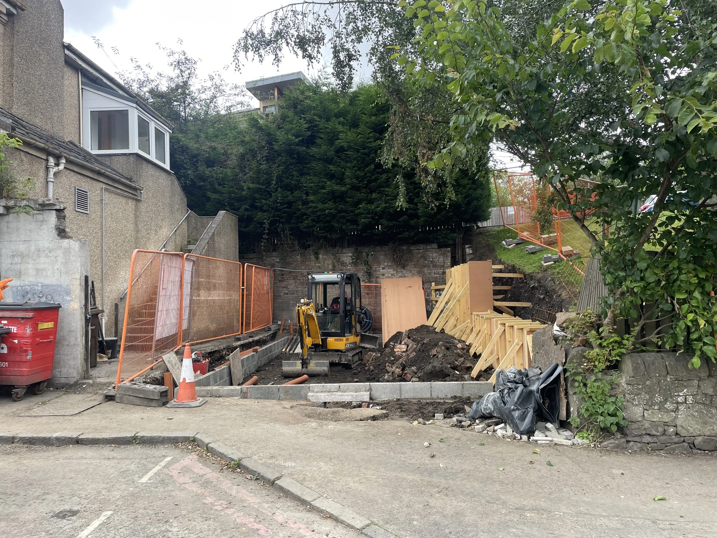 Construction site with a small excavator working on a pathway, surrounded by orange safety barriers, trees, and building walls.