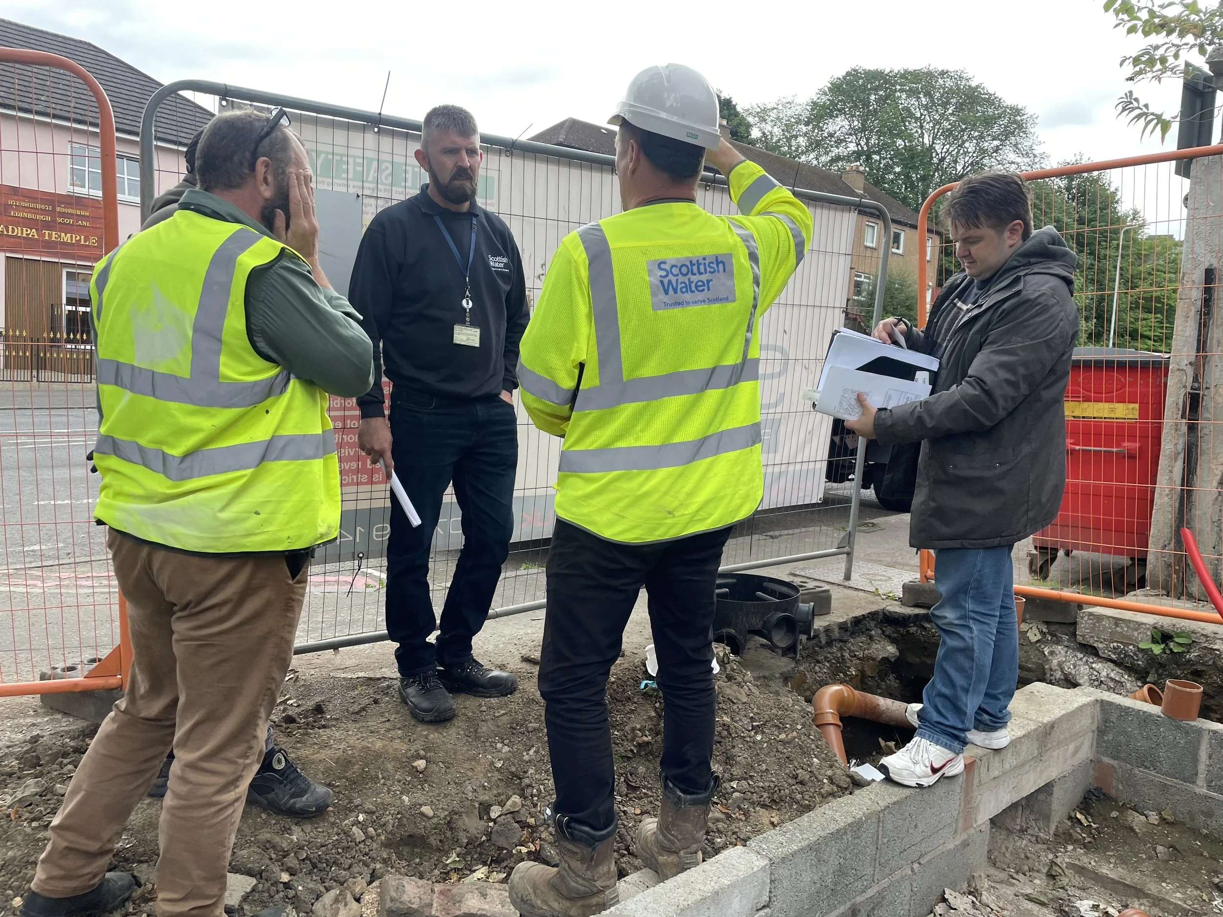 Group of construction workers and officials speaking at a construction site, with orange safety fences and building materials around.