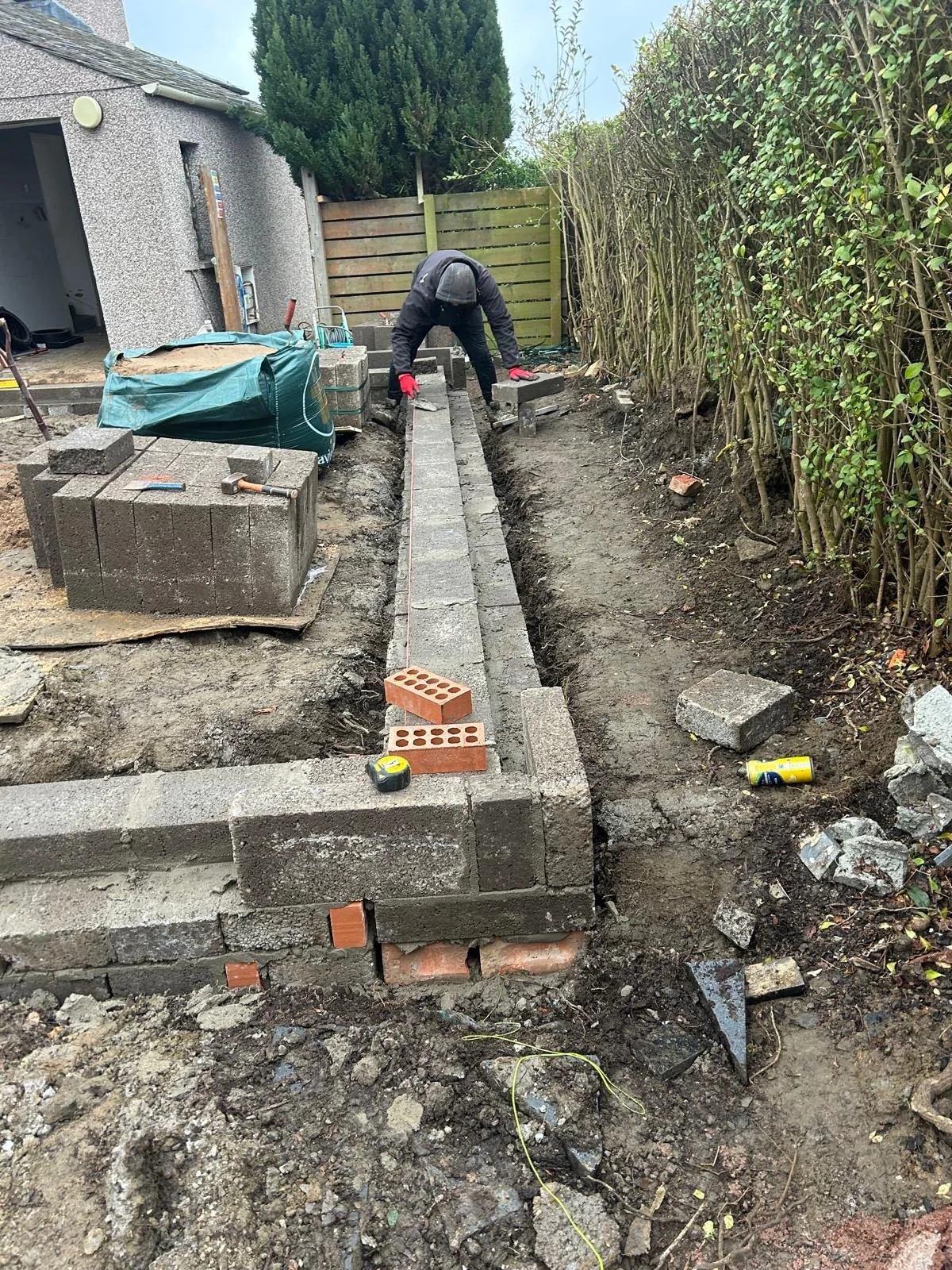 A person is working on constructing a brick wall outdoors, with tools and bricks around them, next to a house and a hedge.