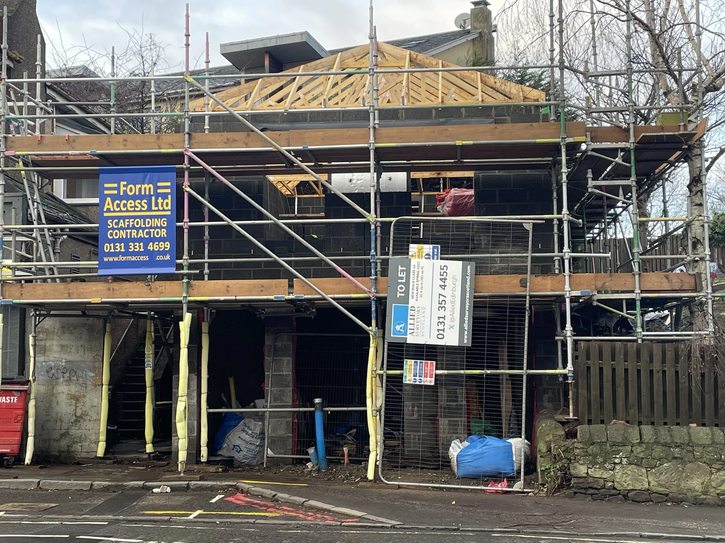 Construction site with scaffolding around a house, showing wooden roof framing, black brick walls, and construction signs.