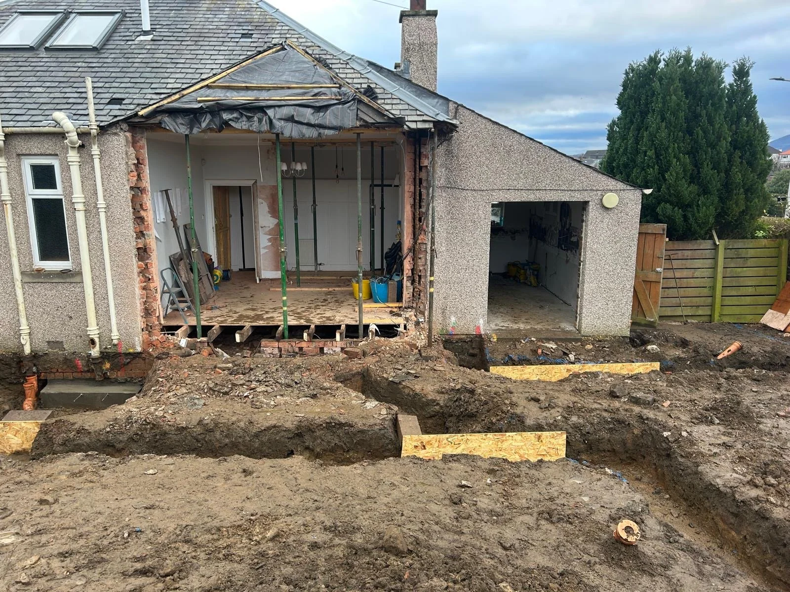 A house undergoing renovation with the exterior wall partially demolished, exposing wooden and brick framework. The front yard is dug up with trenches and dirt, indicating ongoing construction work.