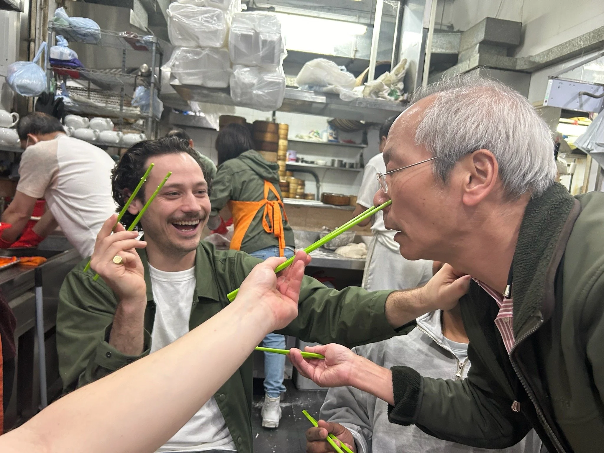Two men are sitting in a restaurant, laughing and using chopsticks, with other people and shelves of dishes and kitchenware in the background.