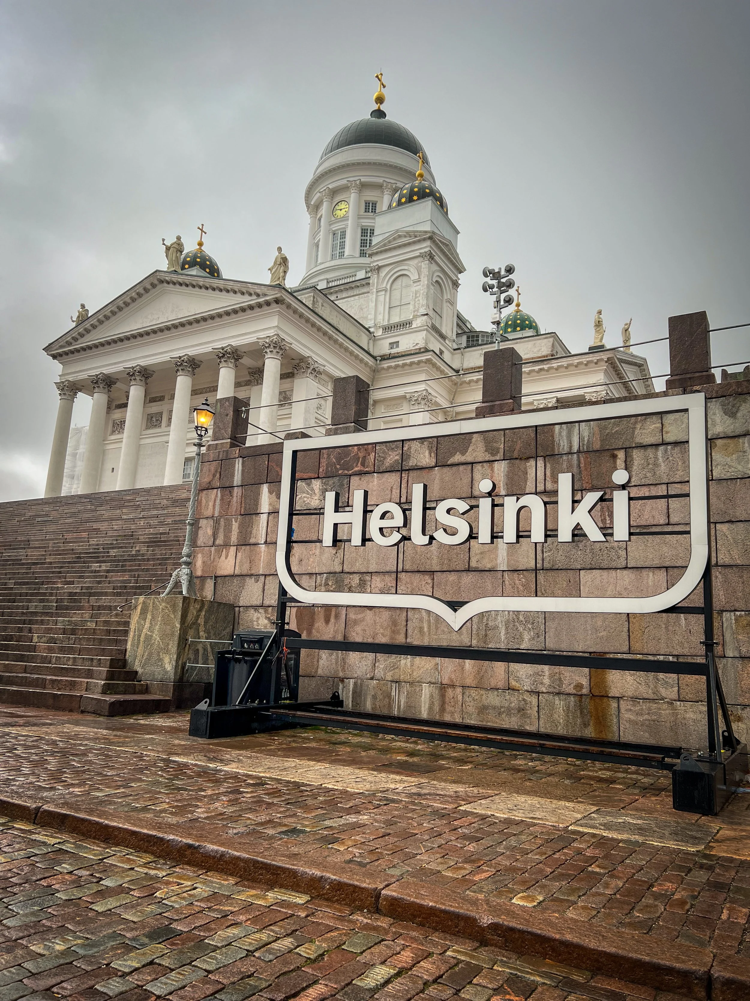 Blick auf die Domkirche in Helsinki mit Treppen, Laterne und einem Schild mit dem Text 'Helsinki' im Vordergrund. Die Kirche hat eine neoklassizistische Architektur mit Säulen und goldenen Heiligenfiguren auf dem Dach. Der Himmel ist bewölkt.